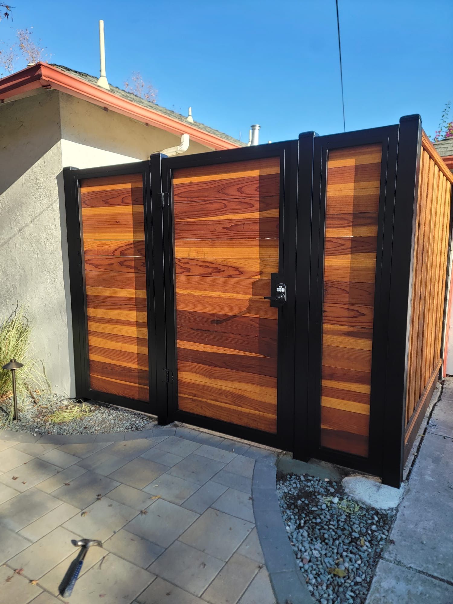A modern fence gate with horizontal wood planks framed in black metal stands on a paved patio beside a house.