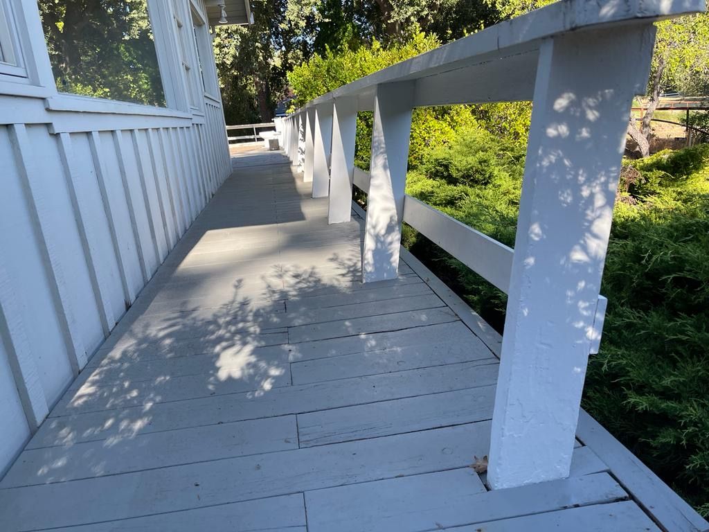 Gray wooden deck with white railing next to a white house, surrounded by trees.