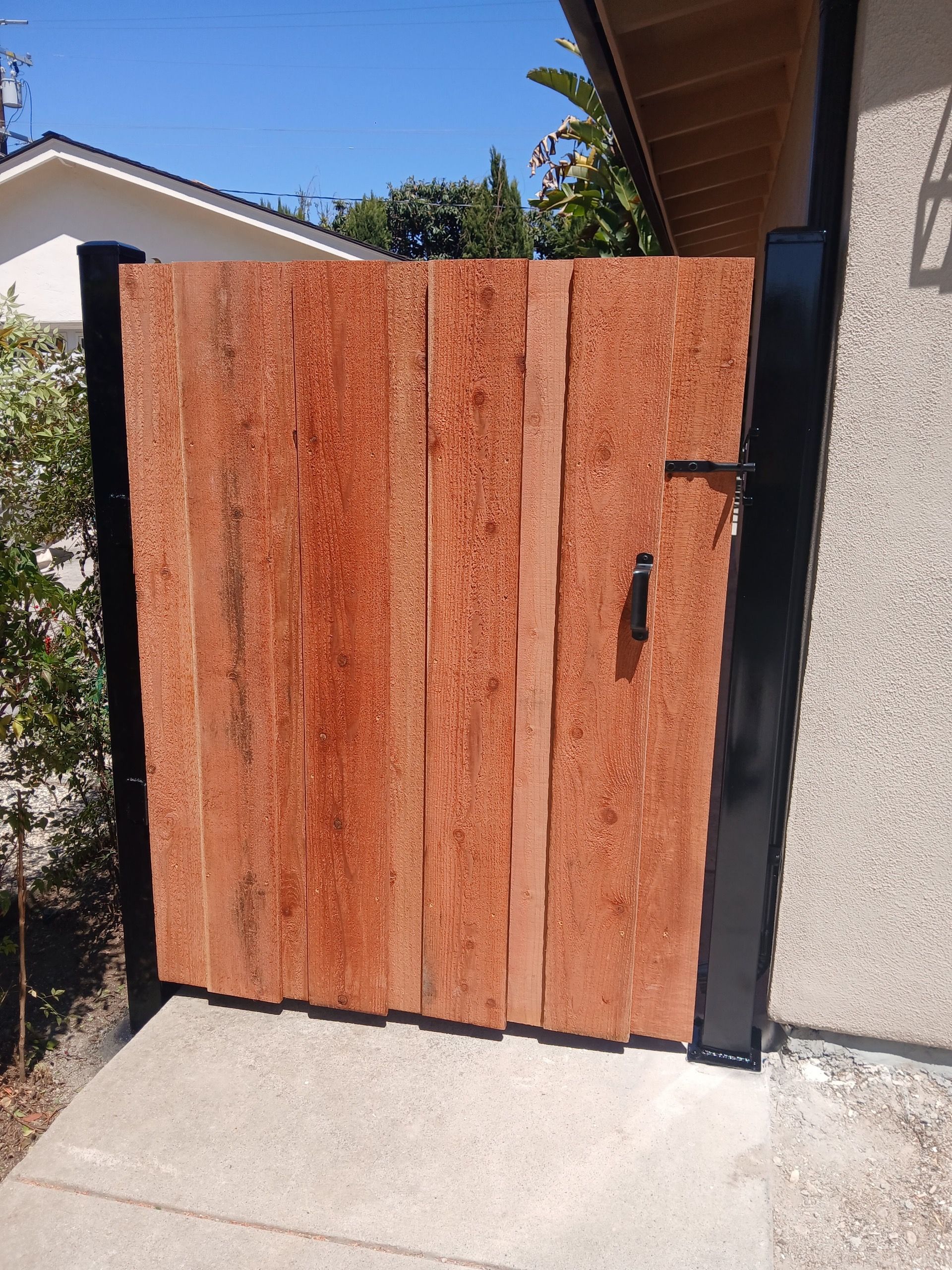 A wooden gate with vertical planks, dark metal frame, and a black latch, installed between a wall and a post outdoors.