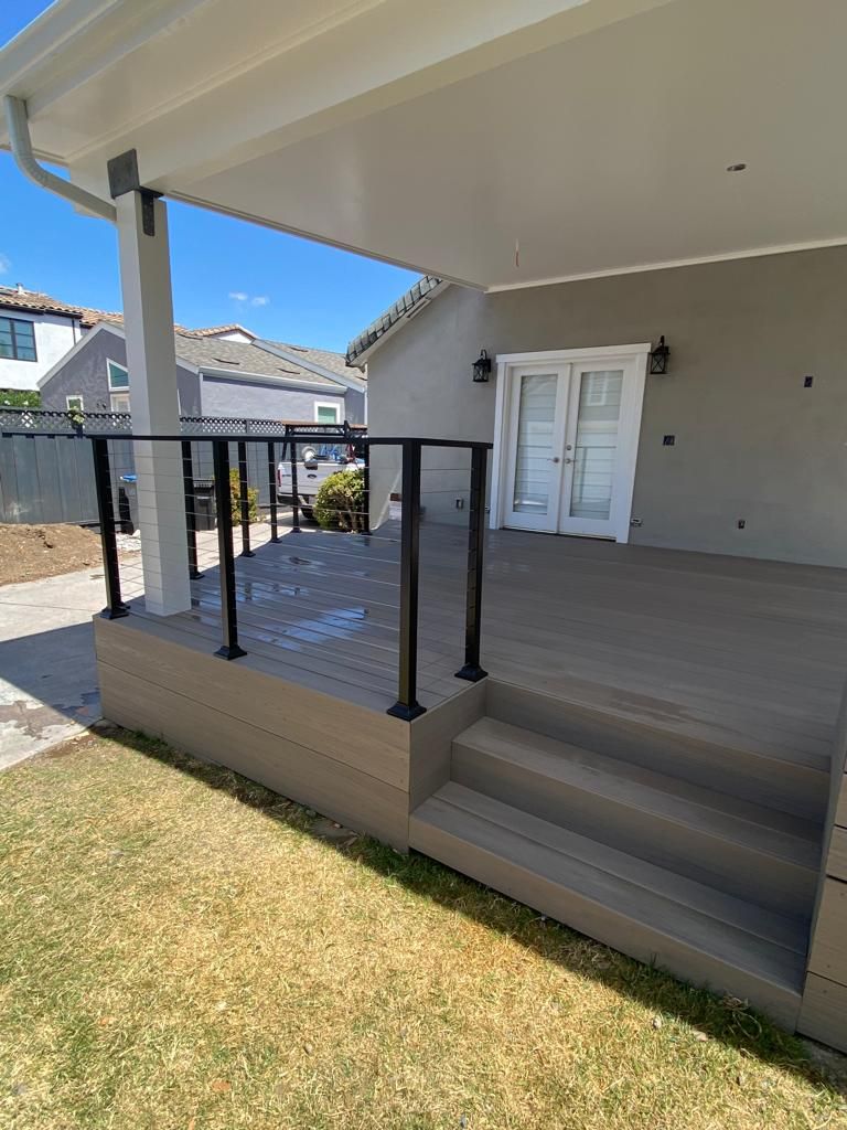 A modern, elevated wooden deck featuring black railings and stairs leading down to a dry, grassy yard under a bright sky.
