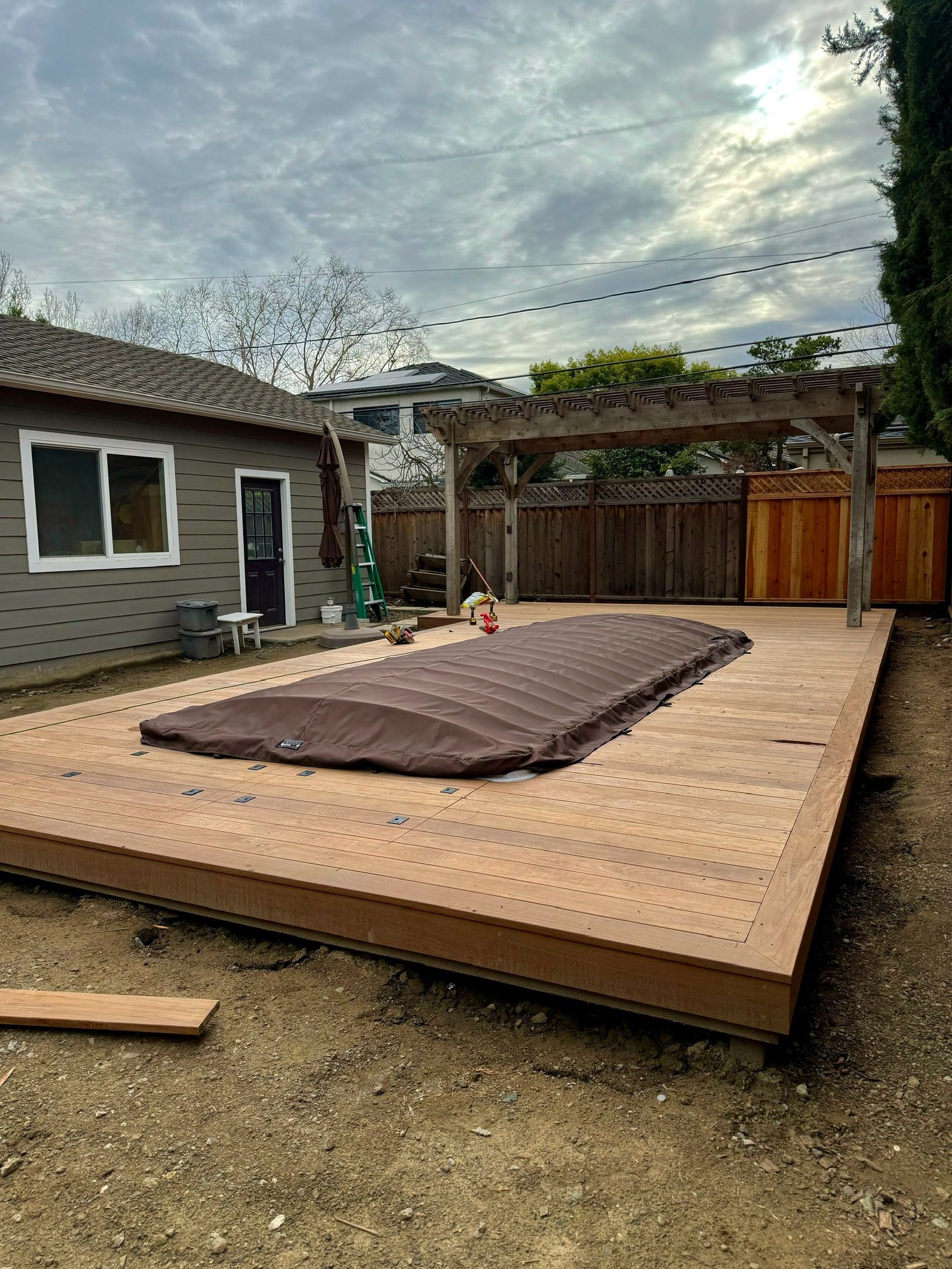 Wooden deck with brown pool cover, arbor, and small house under cloudy sky.