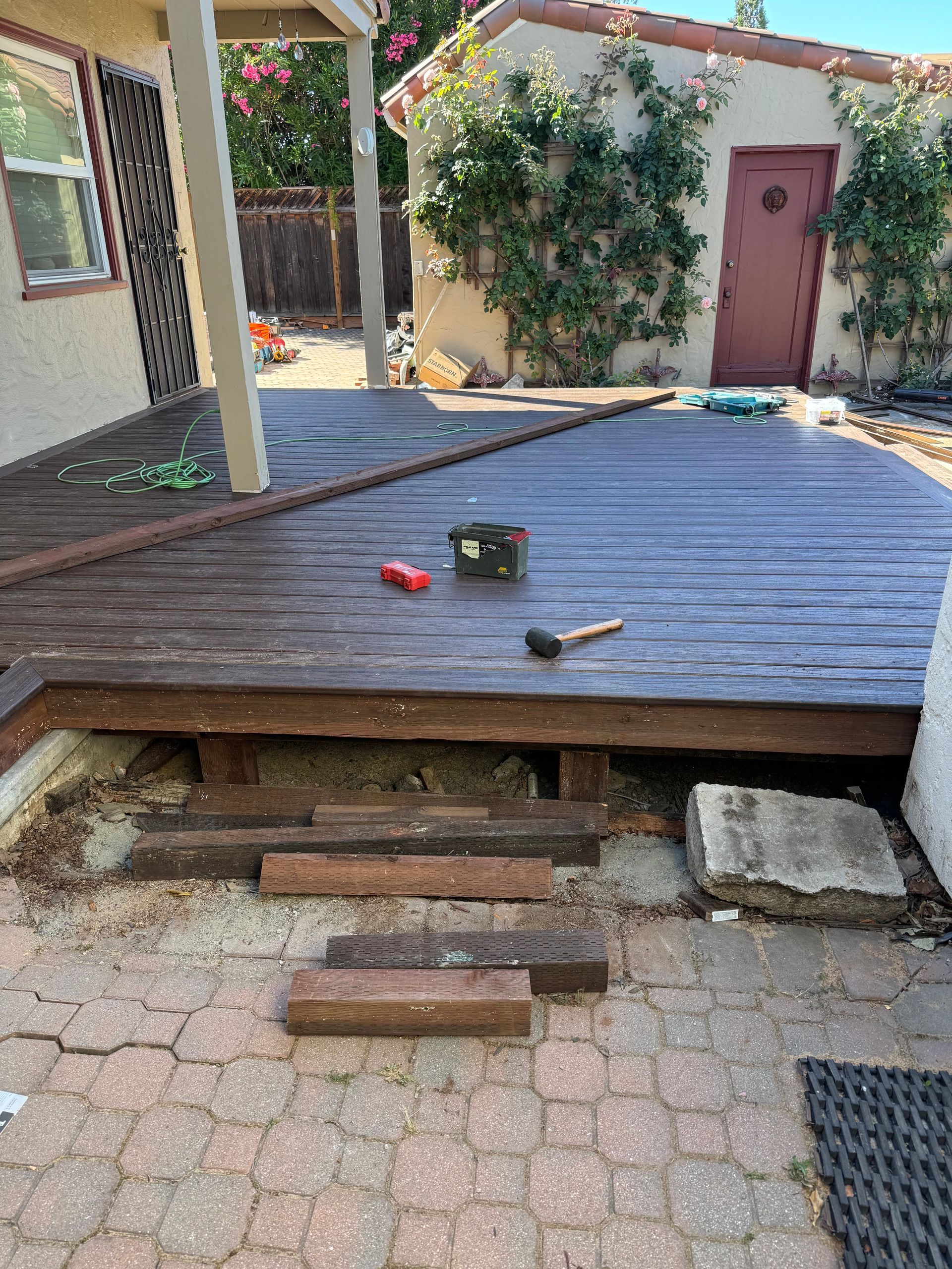 Backyard deck under construction; dark wood planks, brick patio, tools, shed in background.