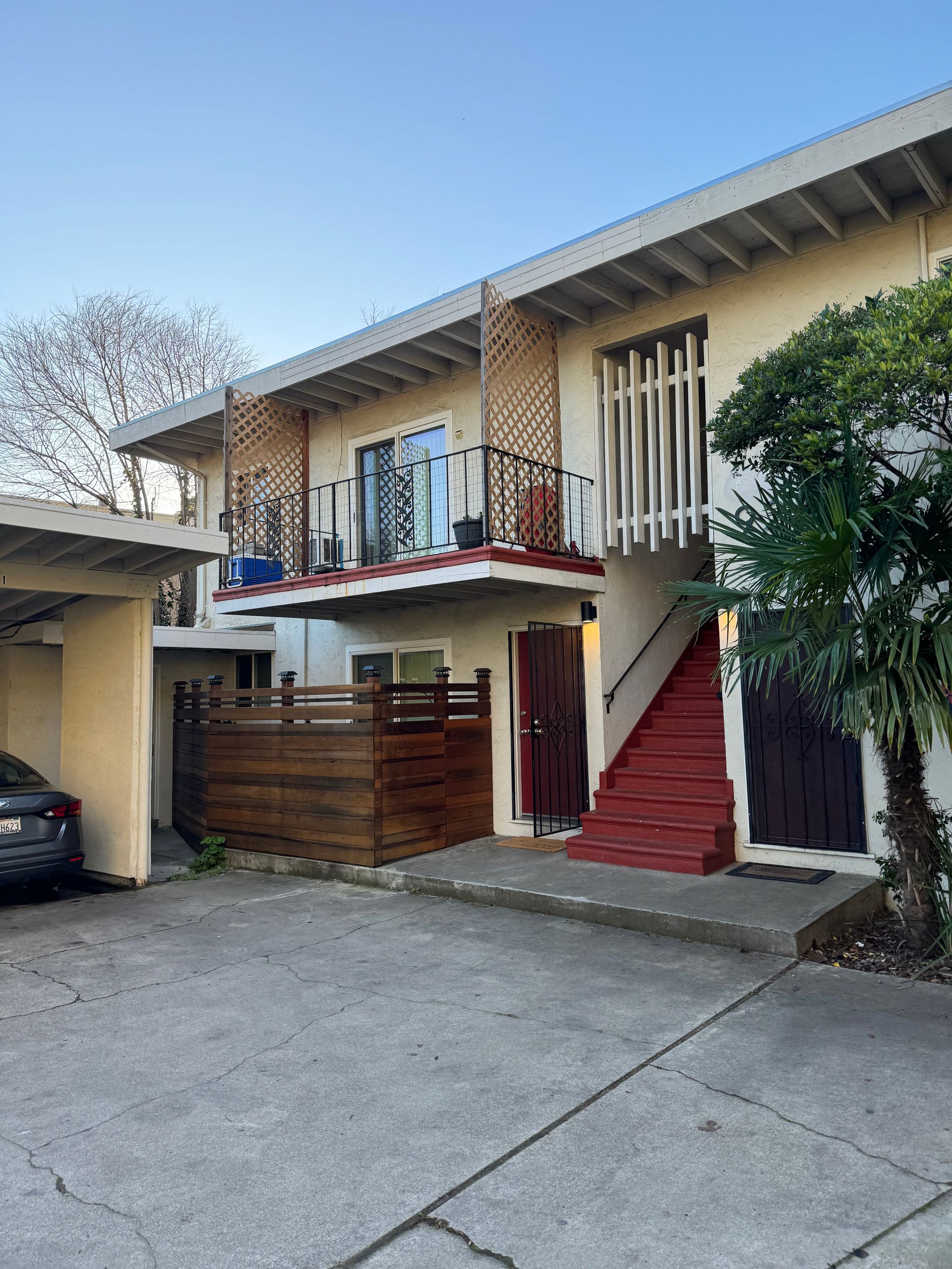 Two-story apartment building with a wooden balcony, red stairs, and a carport on a paved lot under a clear blue sky.