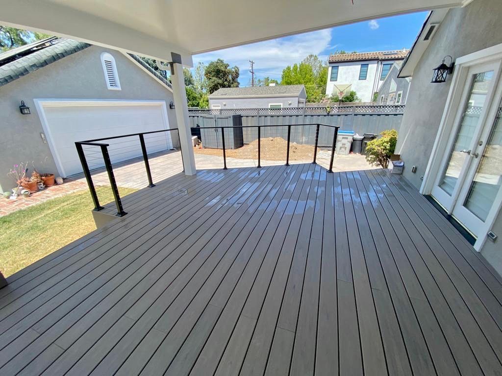 A wide, elevated gray deck with black metal railings, overlooking a backyard with a garage under a bright blue sky.
