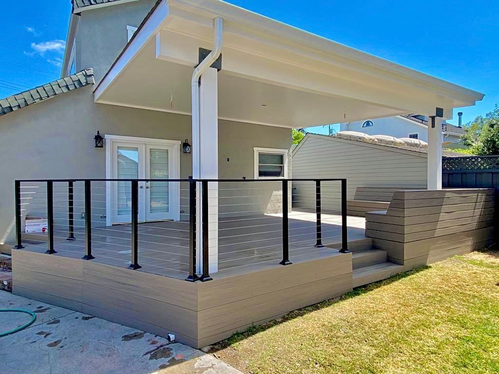 A beige elevated outdoor deck with black railing and a covered patio, featuring a glass door leading into a house.