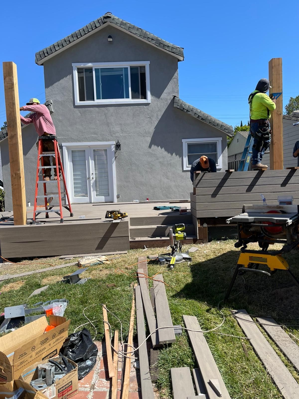Construction workers assemble a deck frame in front of a gray house under a clear blue sky.