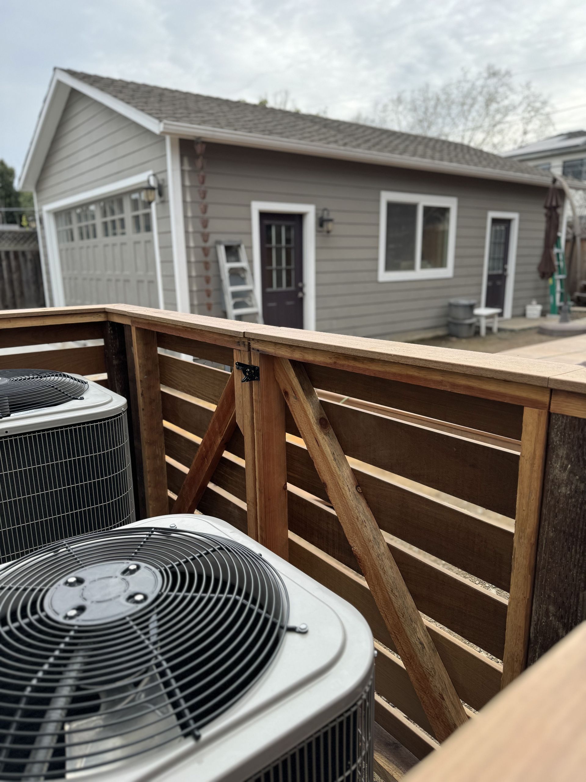 Air conditioning units behind a wooden fence, with a gray garage in the background.