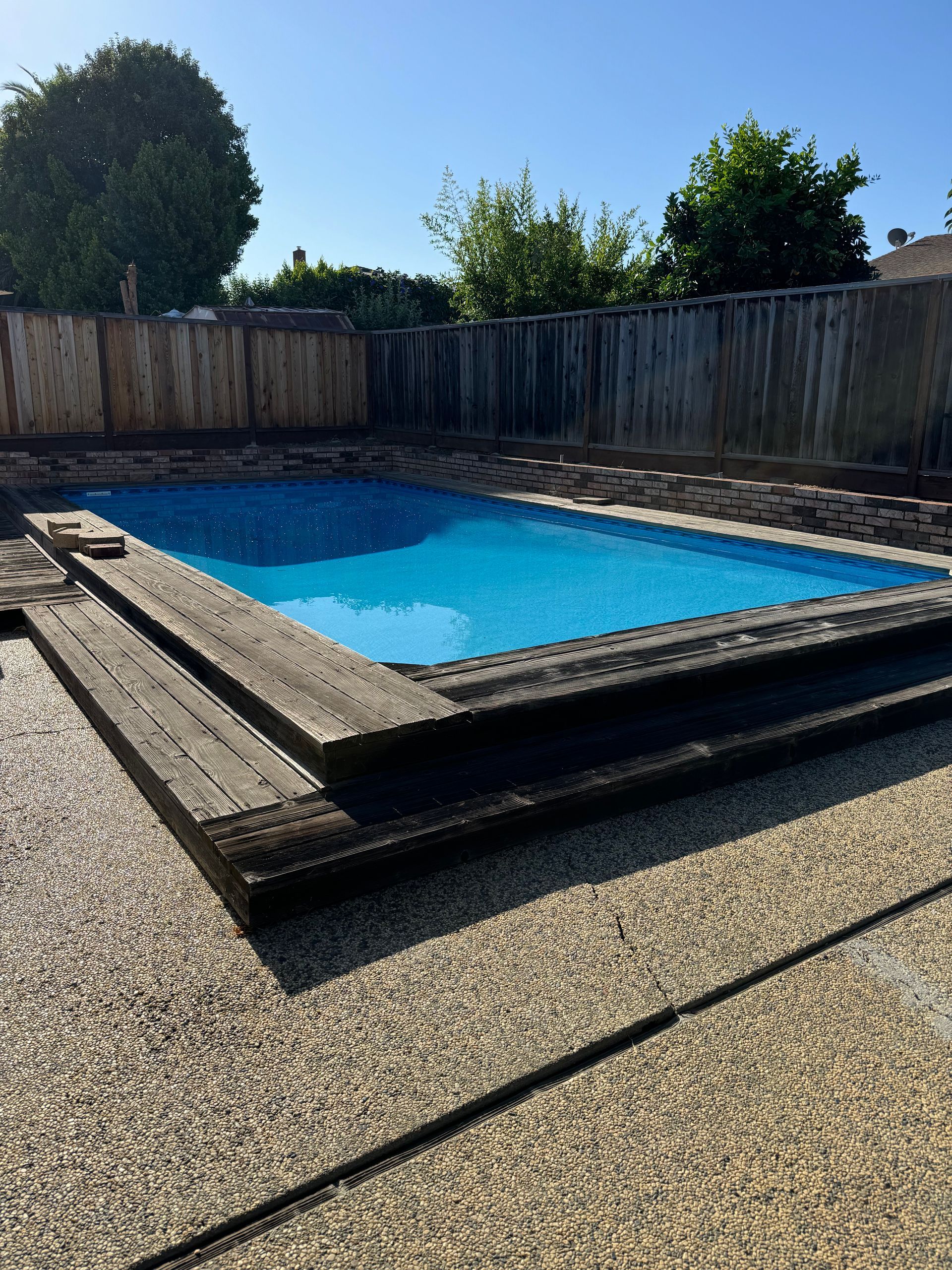 Rectangular pool with blue water, surrounded by wooden frame, gravel, and a wooden fence.