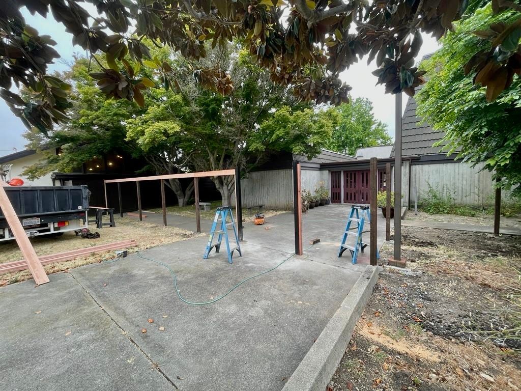 An outdoor patio area under construction, featuring wooden support beams and two blue ladders on a concrete surface.
