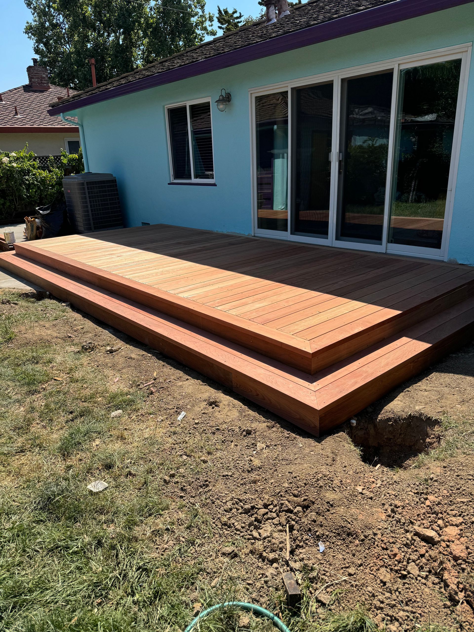 A light blue house features a new, multi-level wooden deck leading to glass sliding doors in a sunny backyard.
