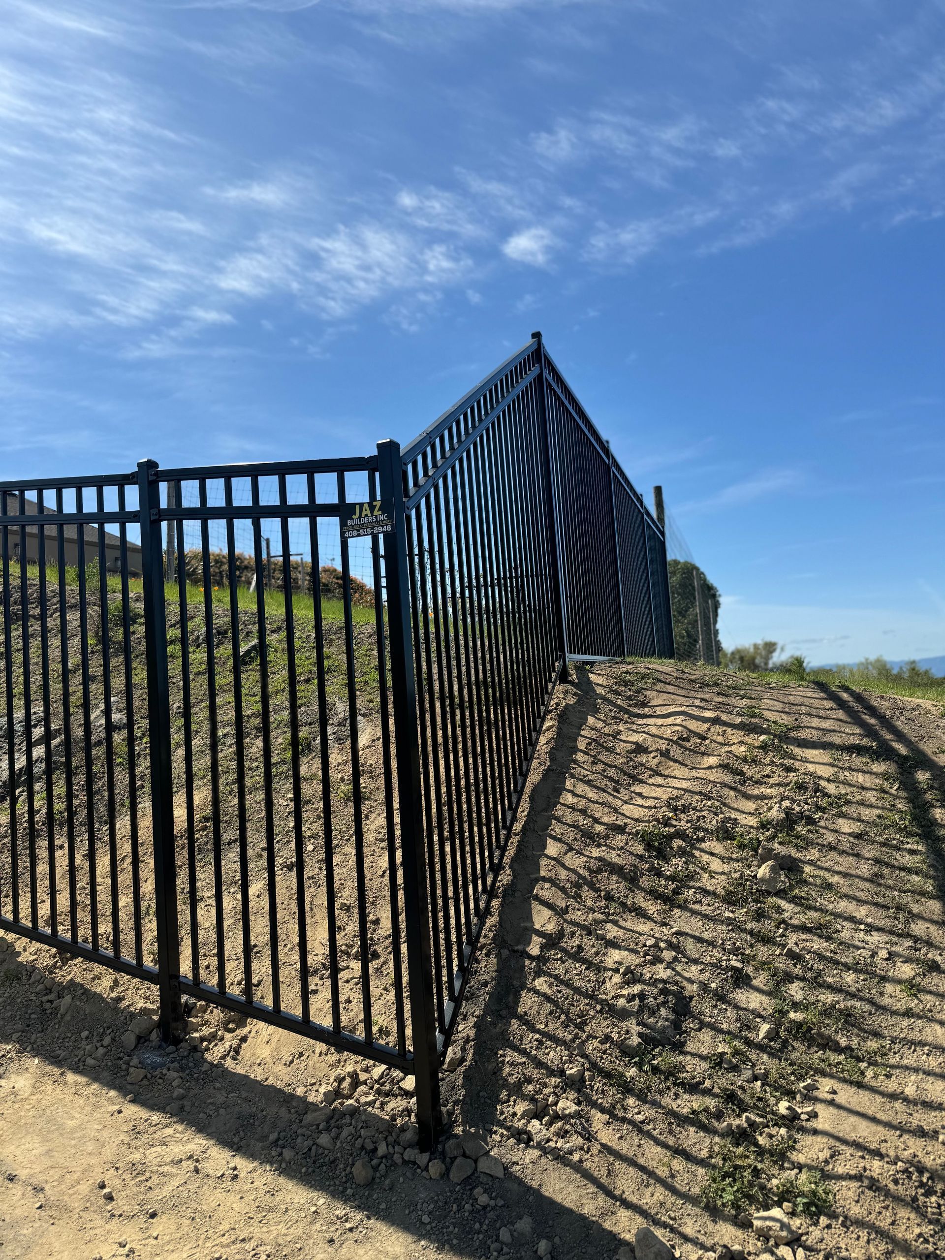 Black metal fence on a hillside under a blue sky, casting a shadow on the dirt.
