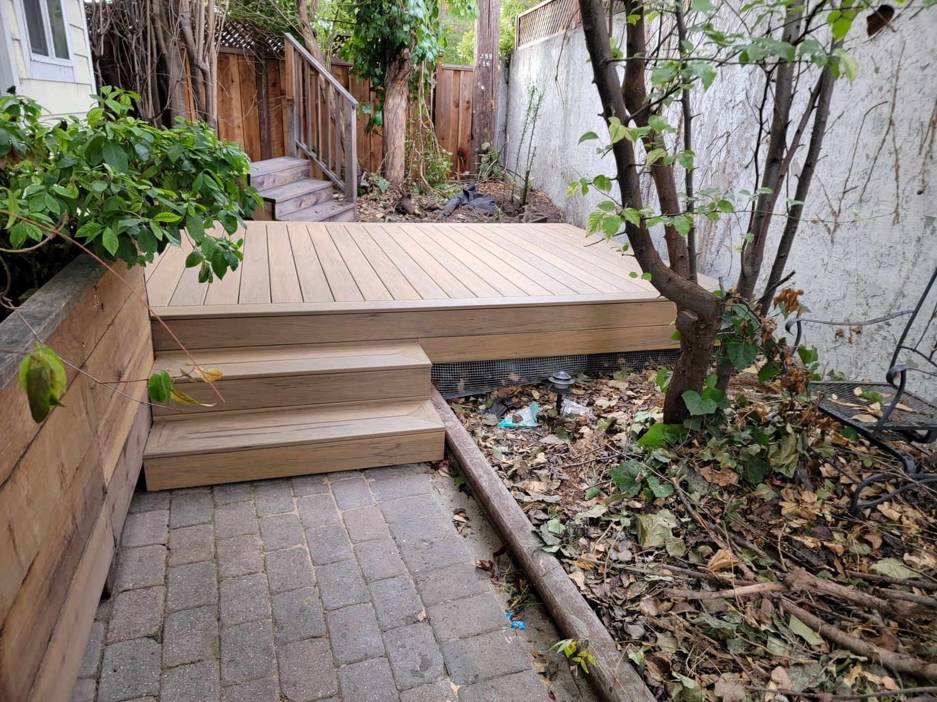 Narrow backyard path with wood steps leading to a raised deck, bordered by trees and a small garden bed