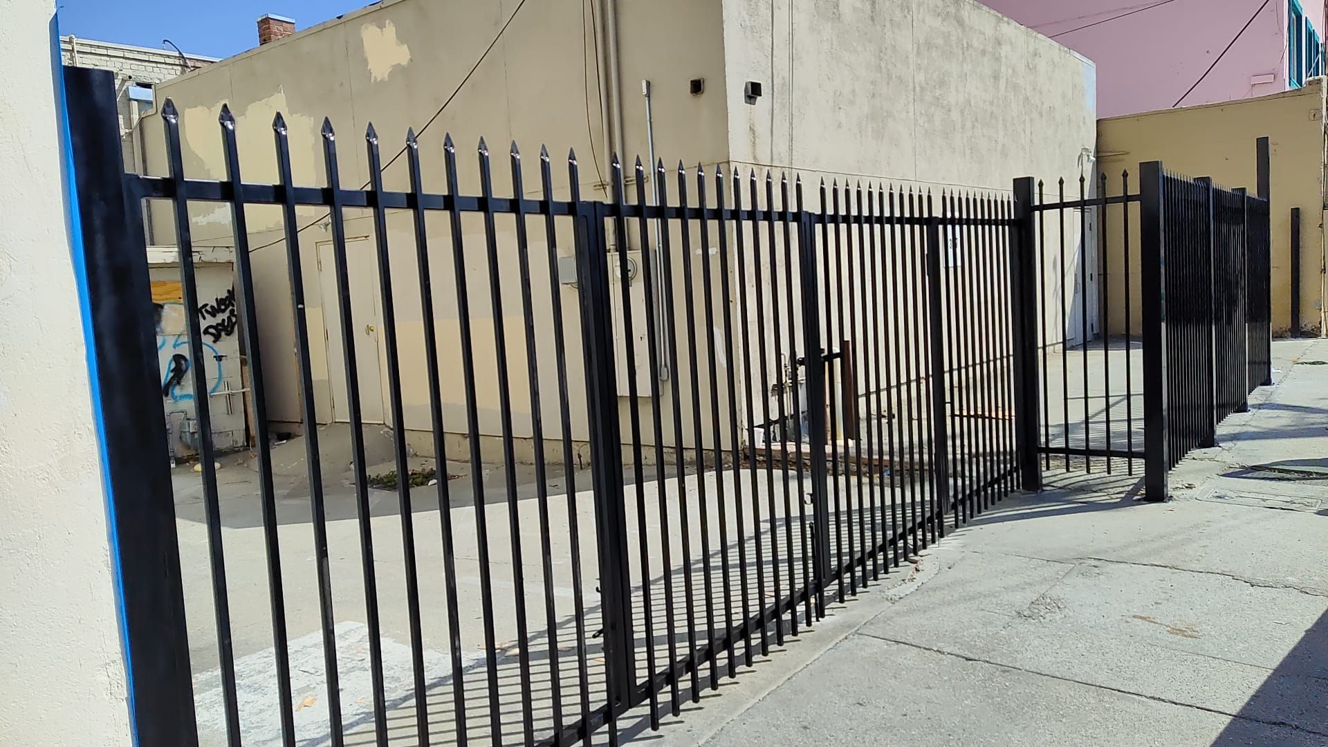 Black metal fence along a sidewalk beside beige buildings on a sunny street.