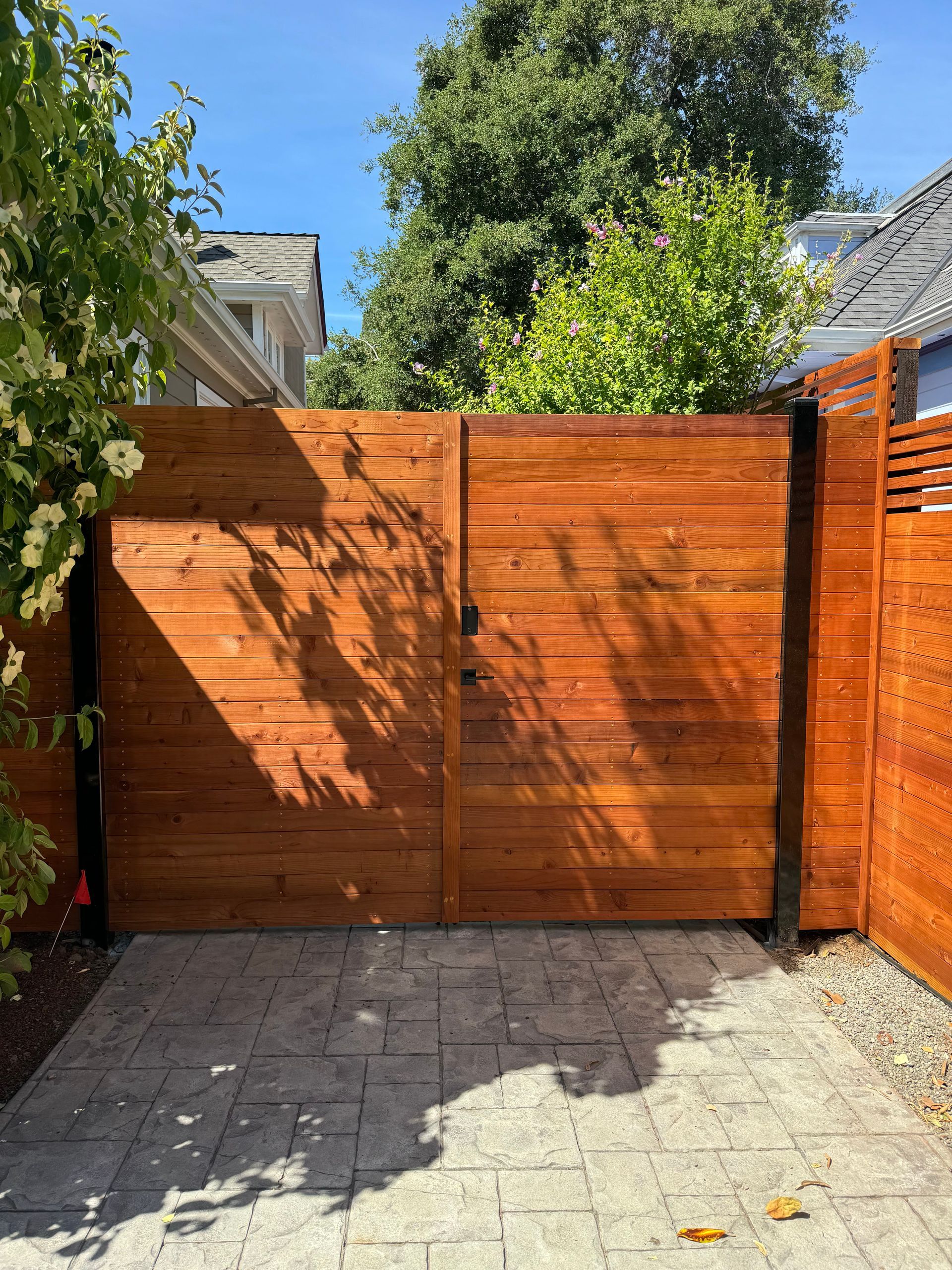 Wooden driveway gate and fence in a sunny backyard, with trees and stone paving.