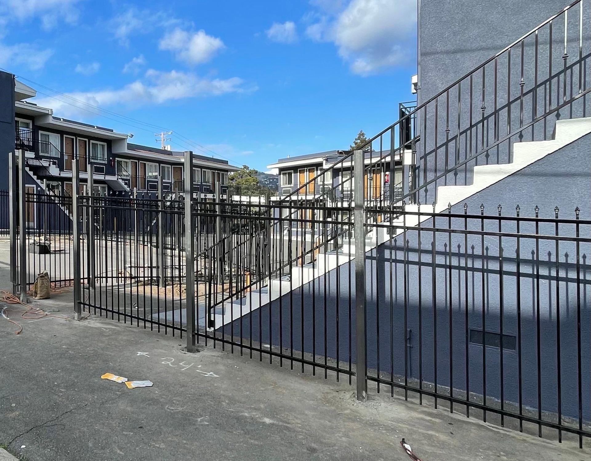 Black metal fence along a gray building with white stairs, leading to other buildings under a blue sky.