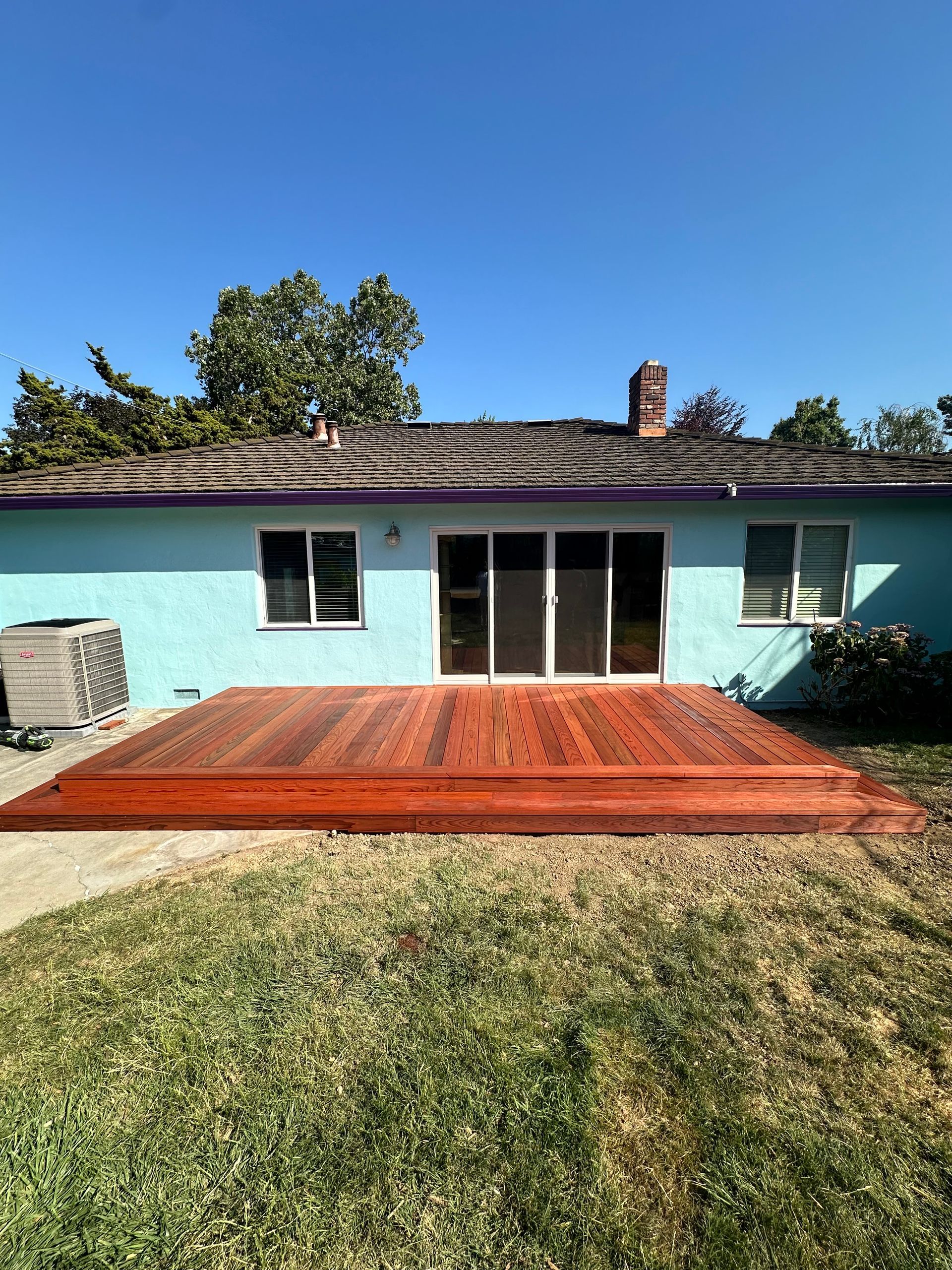A newly built wooden deck extends from the back of a light blue house with a dark roof on a sunny day.