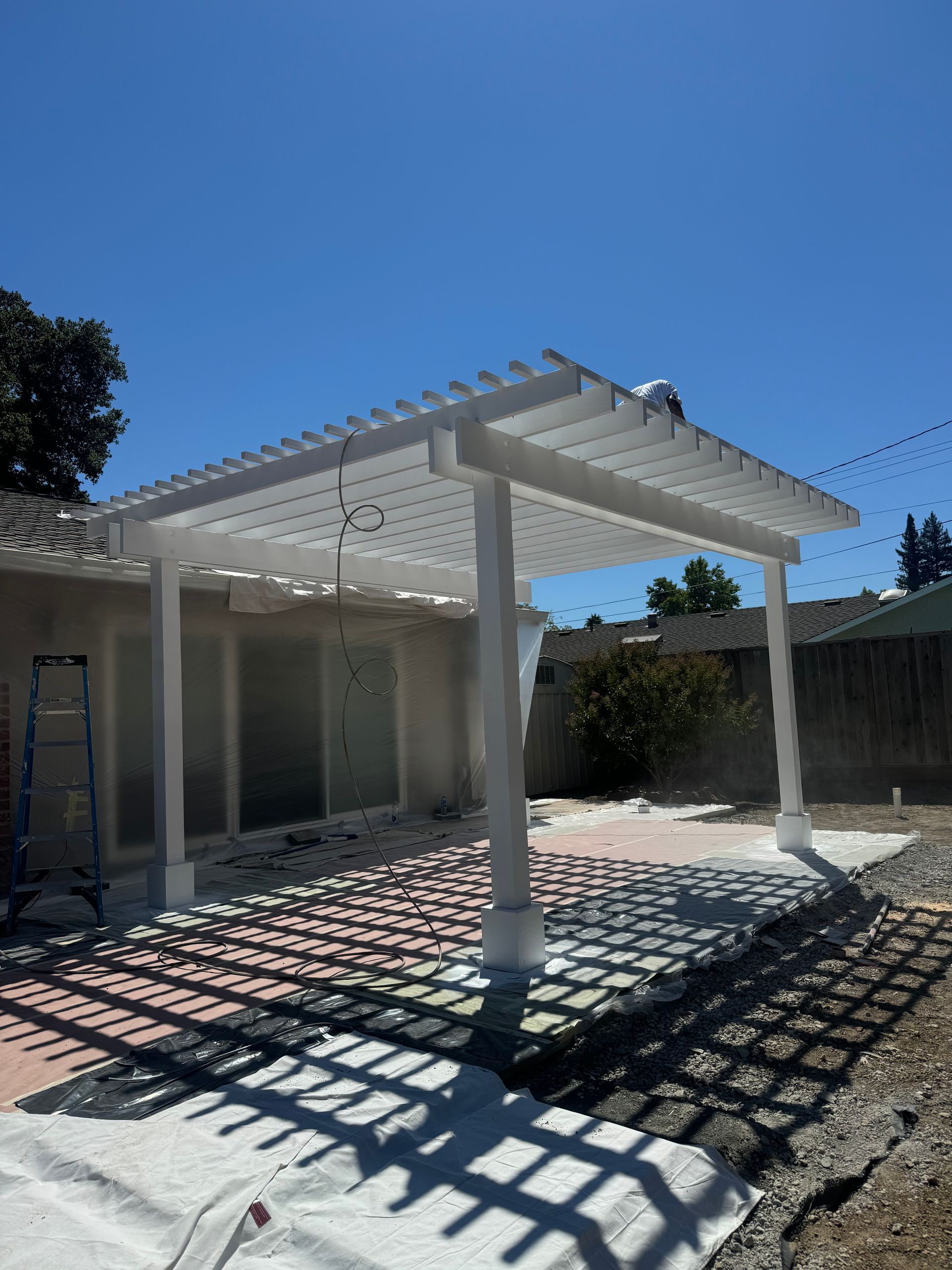 White pergola patio in a sunlit backyard with strong shadow patterns on the ground