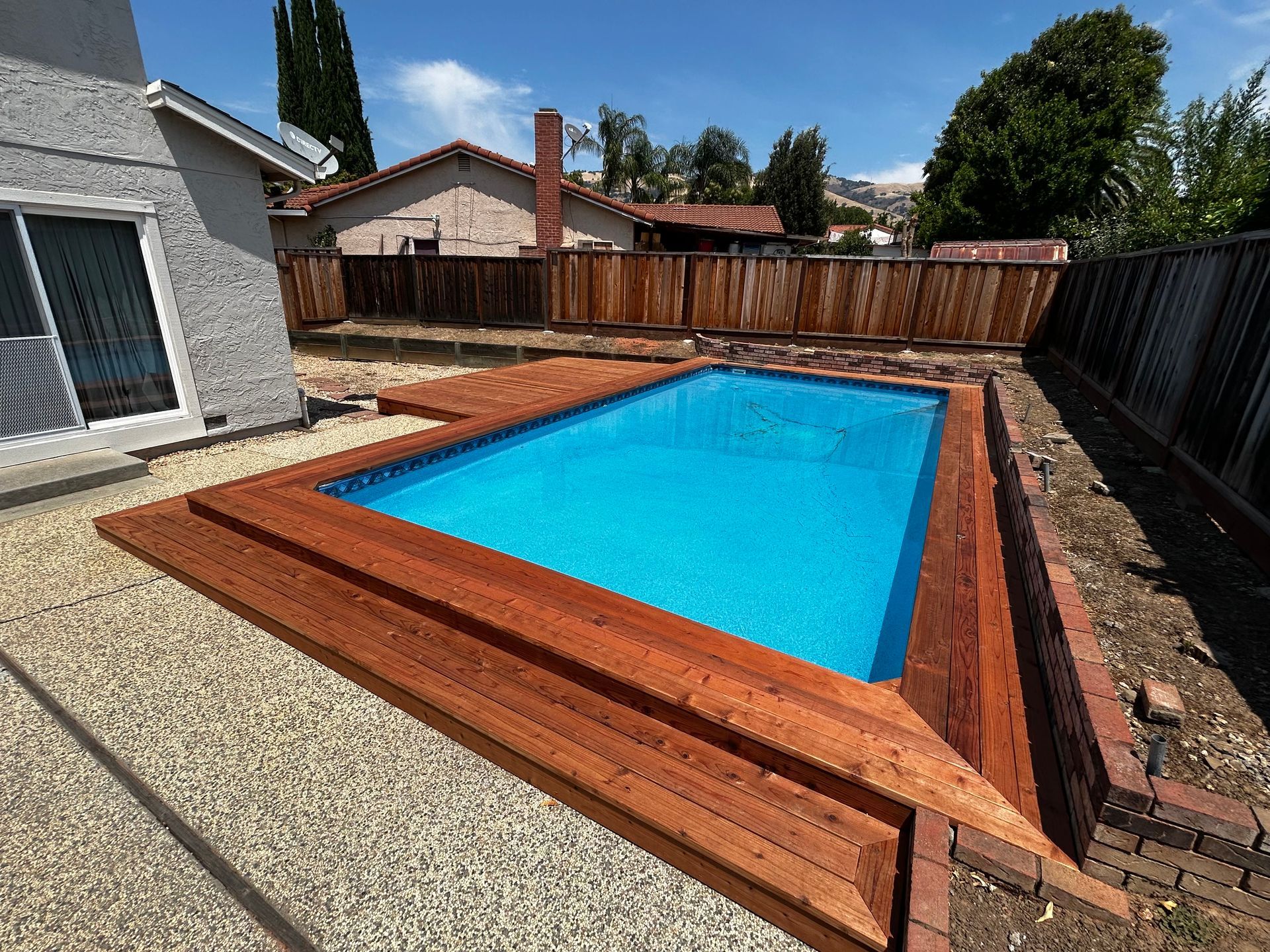 A bright blue rectangular swimming pool with a natural wood deck surround, set in a backyard near a house and fence.
