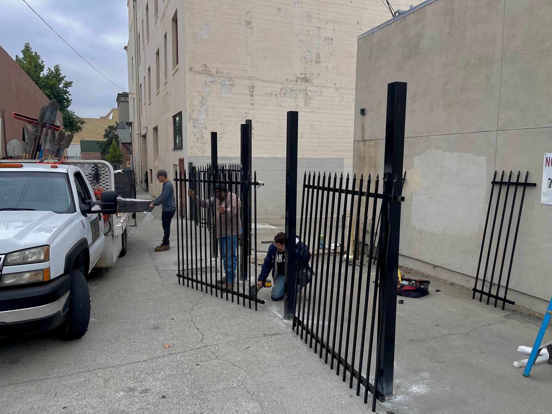 People working beside a black metal fence in a narrow alley next to white buildings and parked trucks.