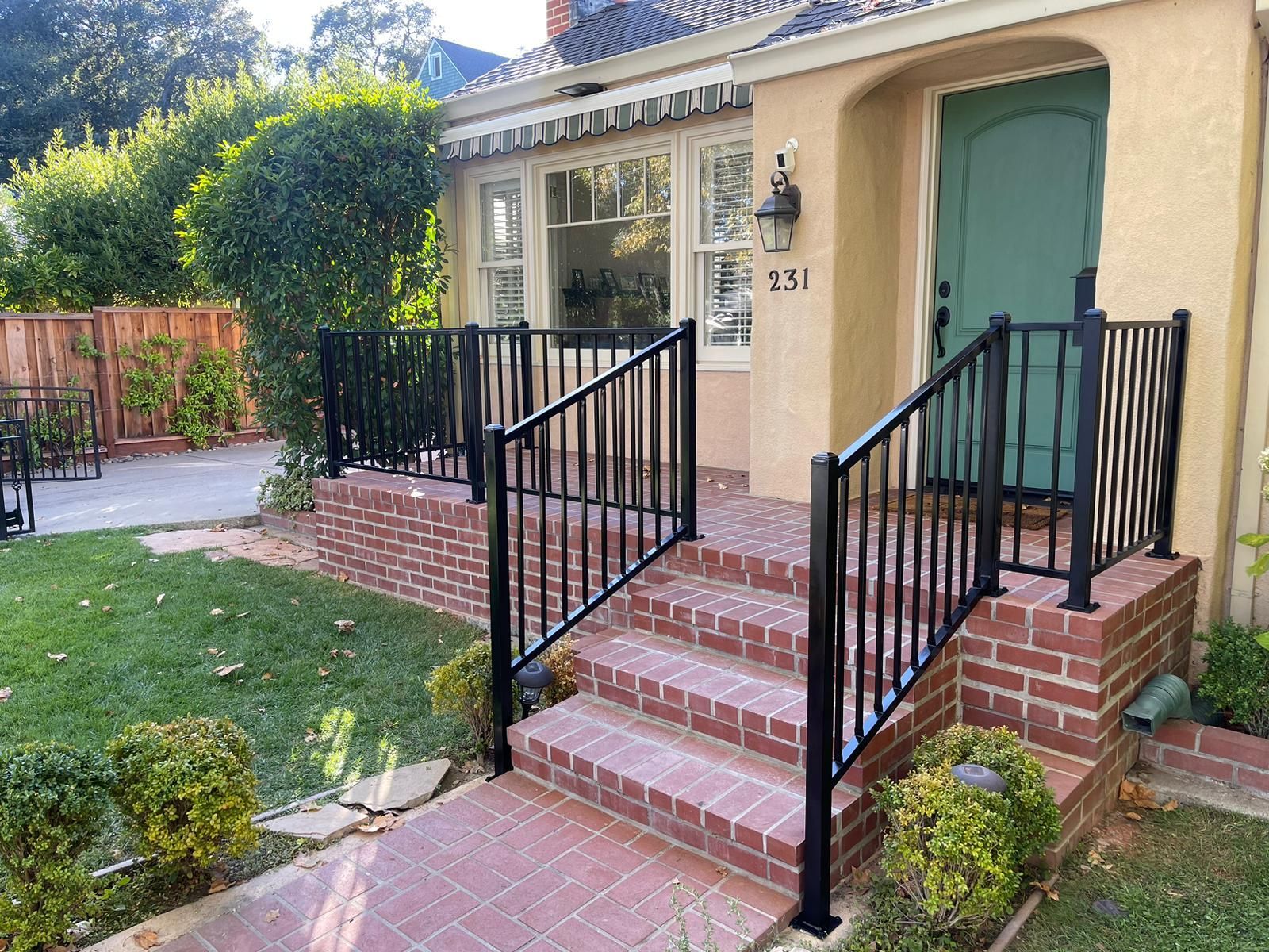 A brick porch with black metal railings leading to a light-colored house with a green door.