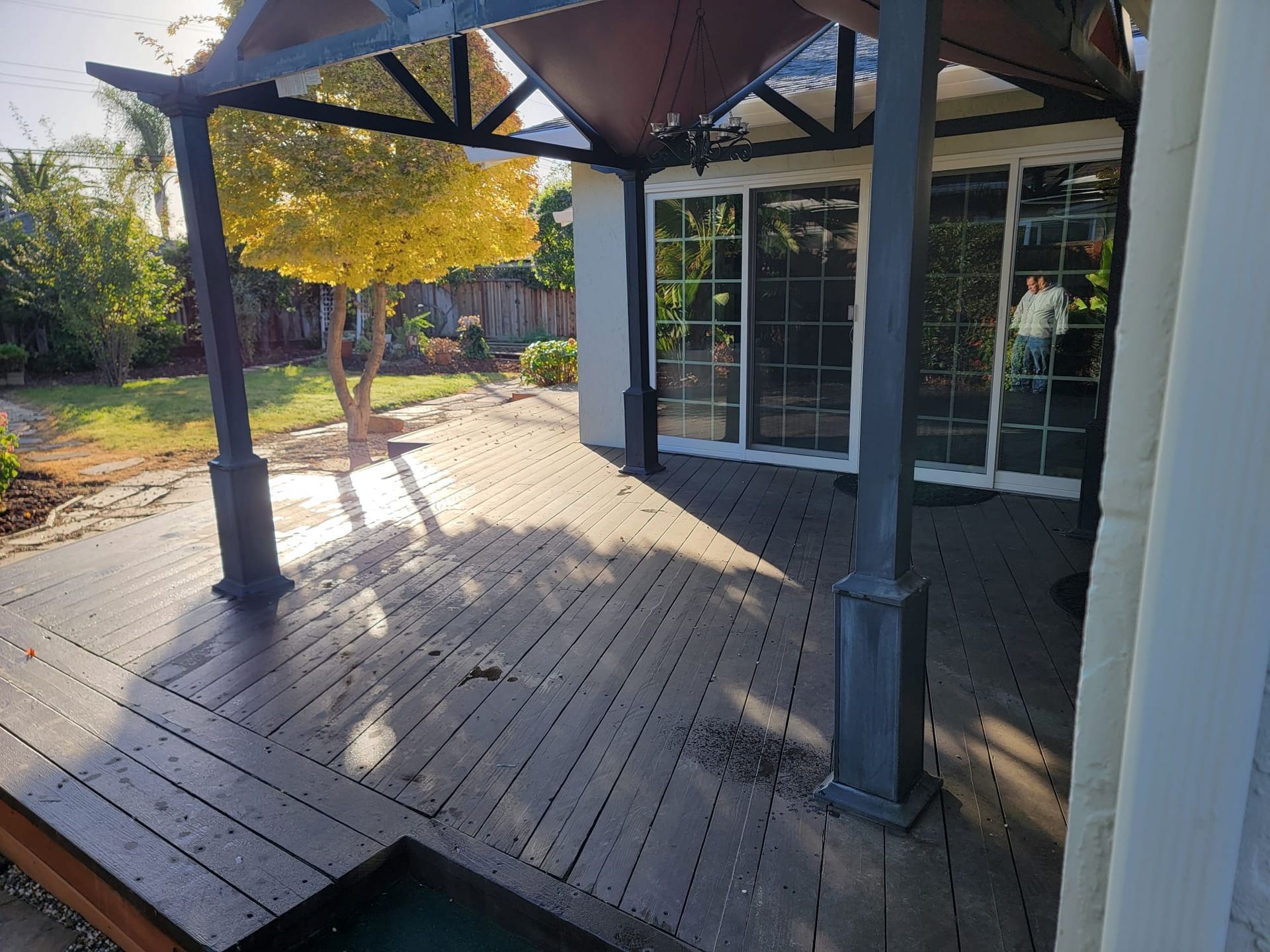 Covered wooden patio with metal columns and glass doors, sunlit yard beyond