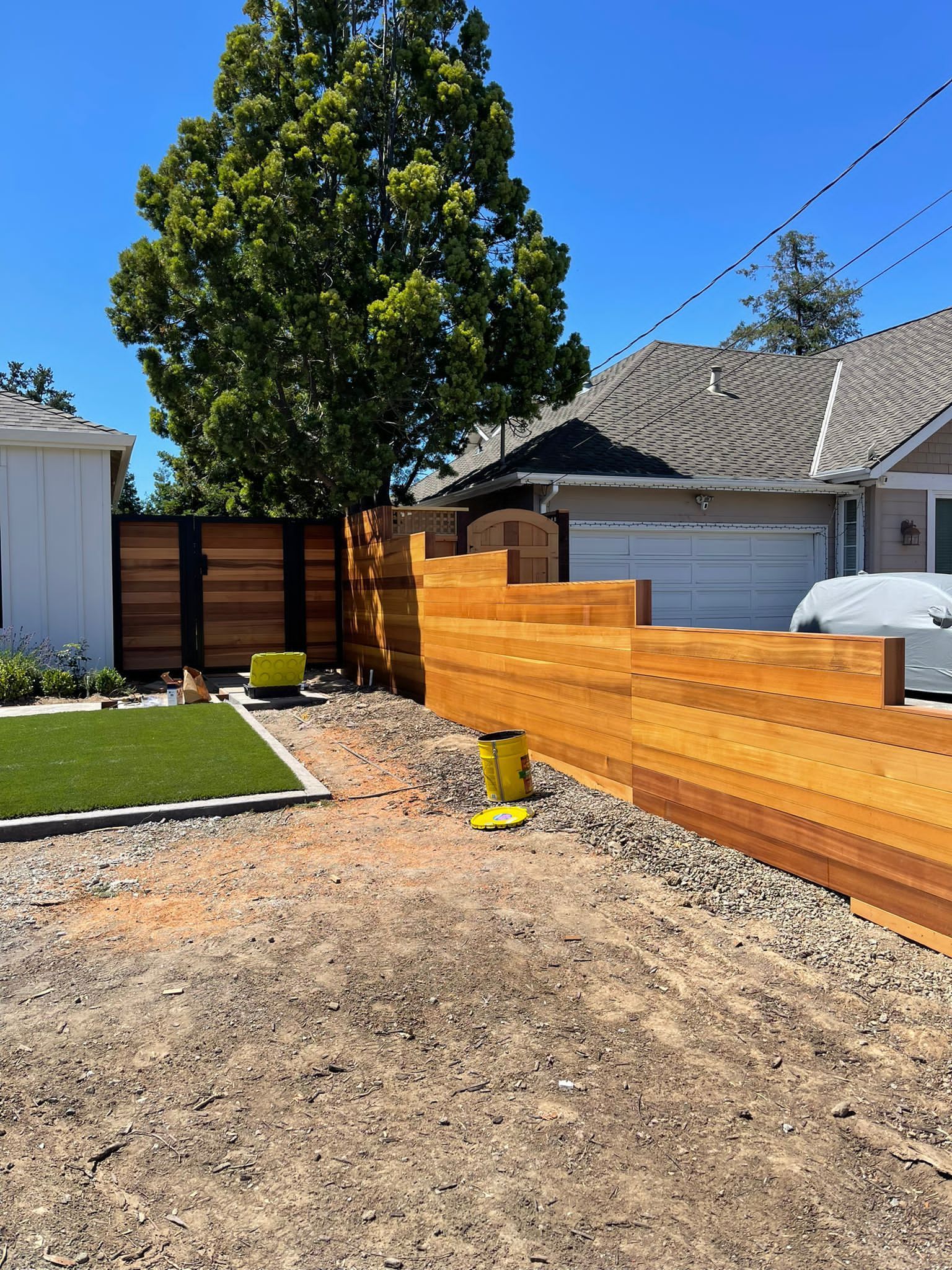A modern horizontal wood fence sits in a yard with gravel landscaping, a grass patch, and a house in the background.