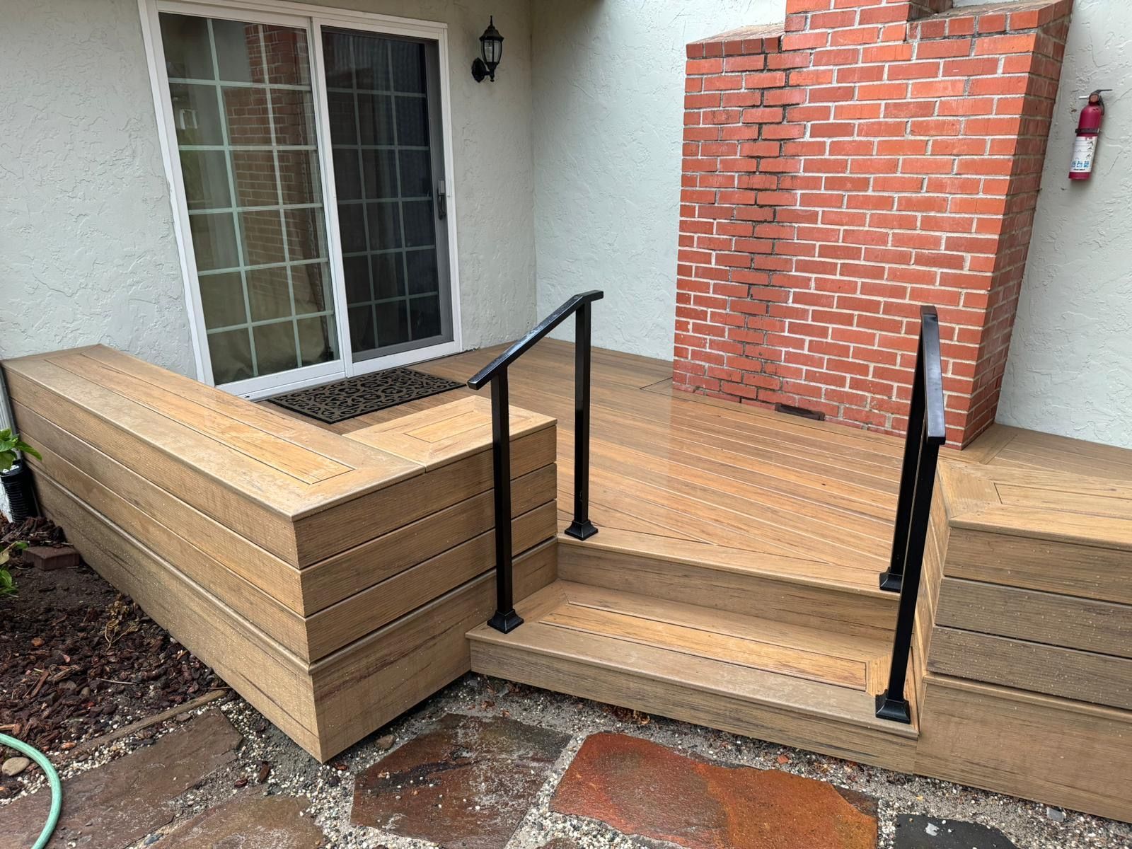A wooden deck with built-in bench seating and a small set of steps with black metal handrails outside a brick chimney.