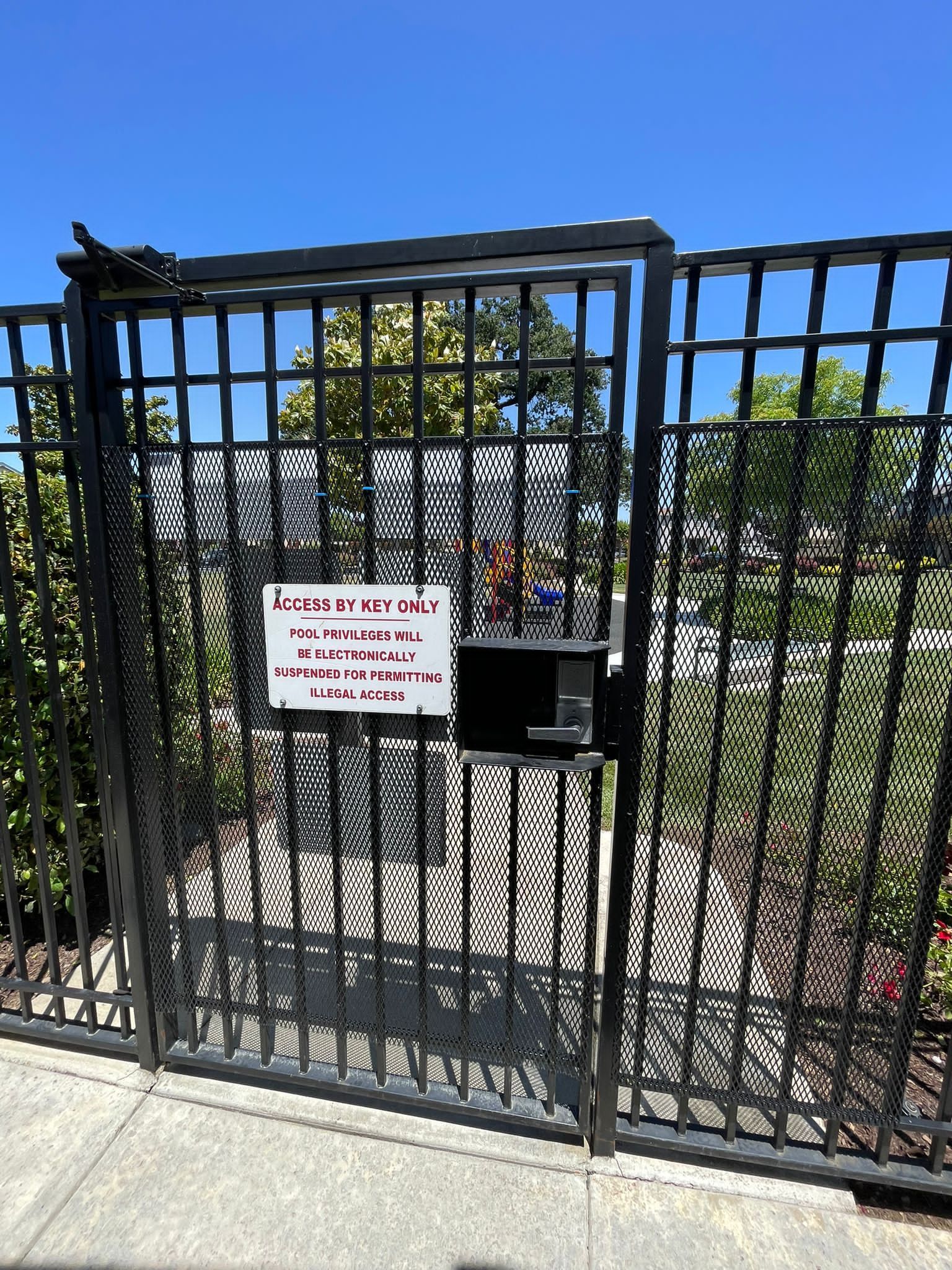 A black metal gate with a keypad lock and a white sign on a sidewalk under a clear blue sky.