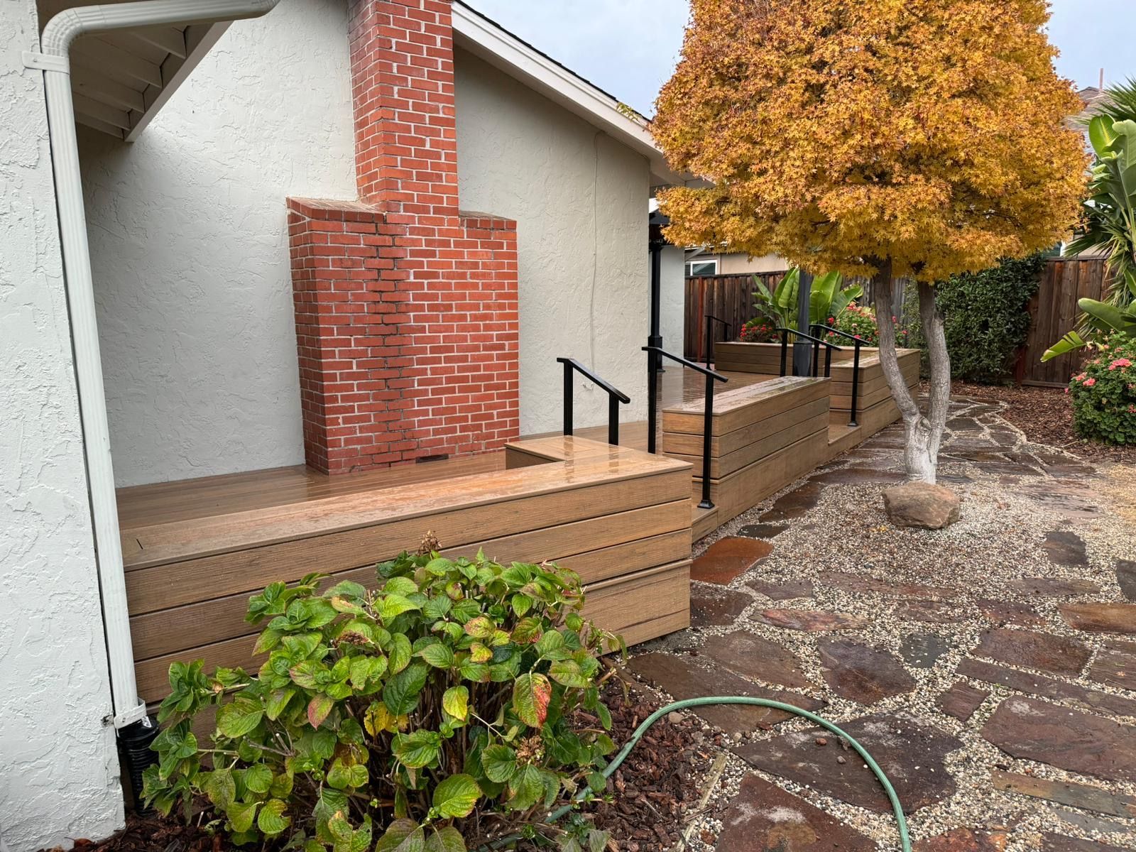 A tan wooden deck with black railings surrounds a red brick chimney on the exterior of a white house beside a stone patio.