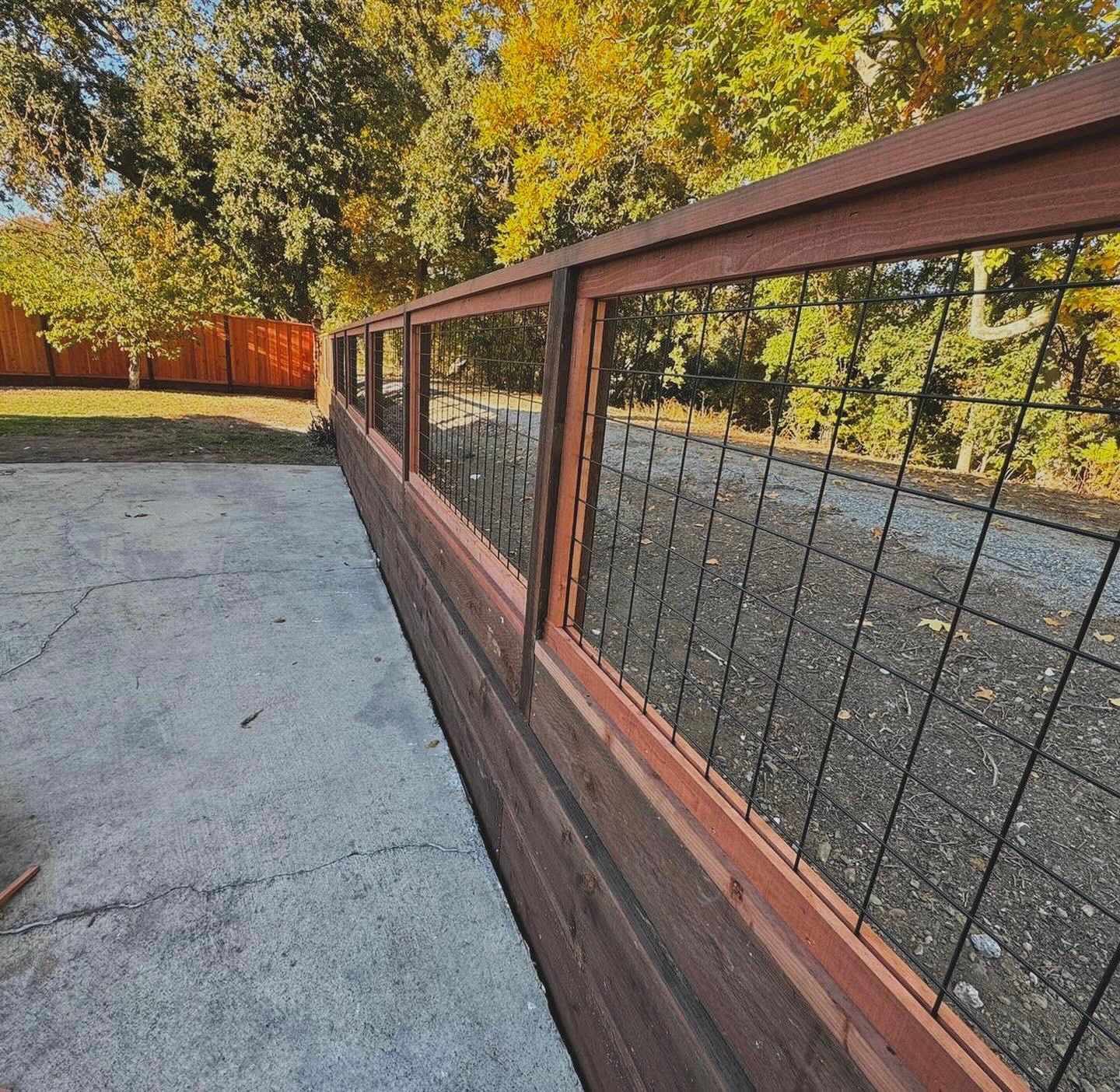 Concrete patio beside a wooden fence with wire panels, bordered by autumn trees and a red privacy fence.