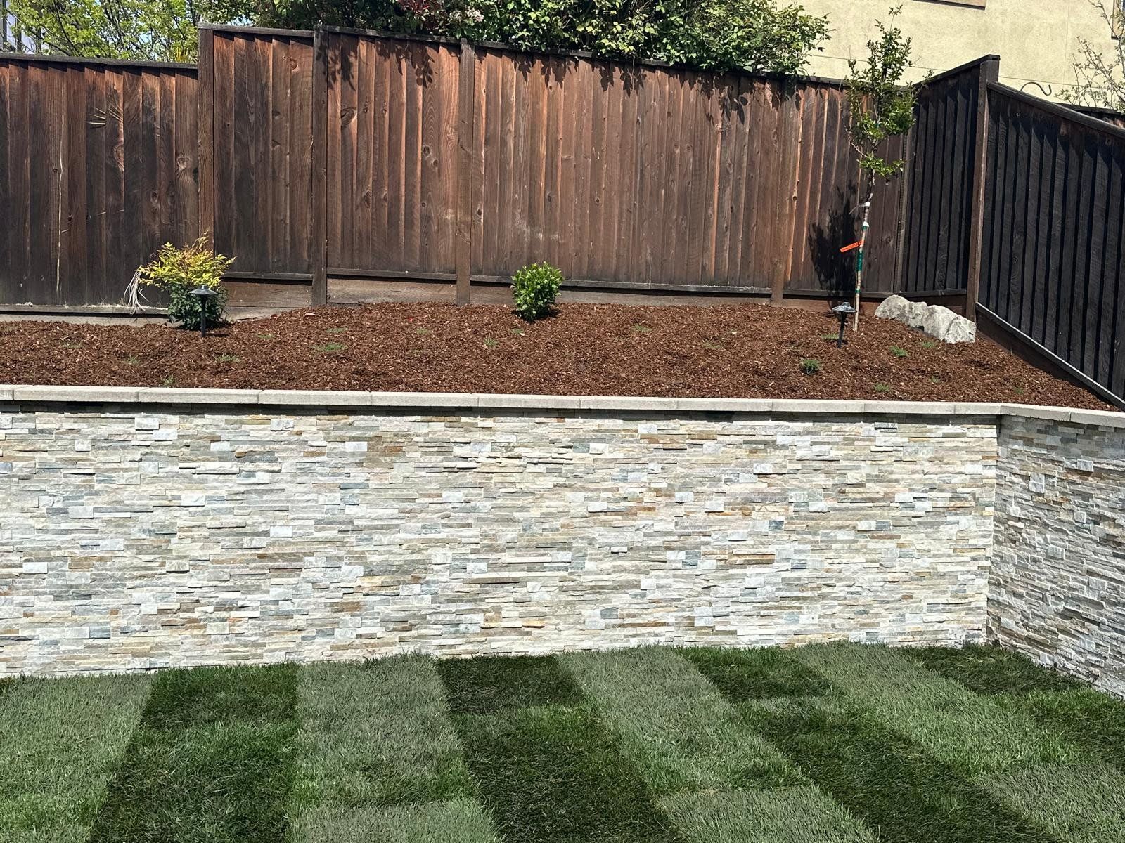 Tiered backyard with wooden fence, stone retaining wall, mulch bed, and striped green lawn