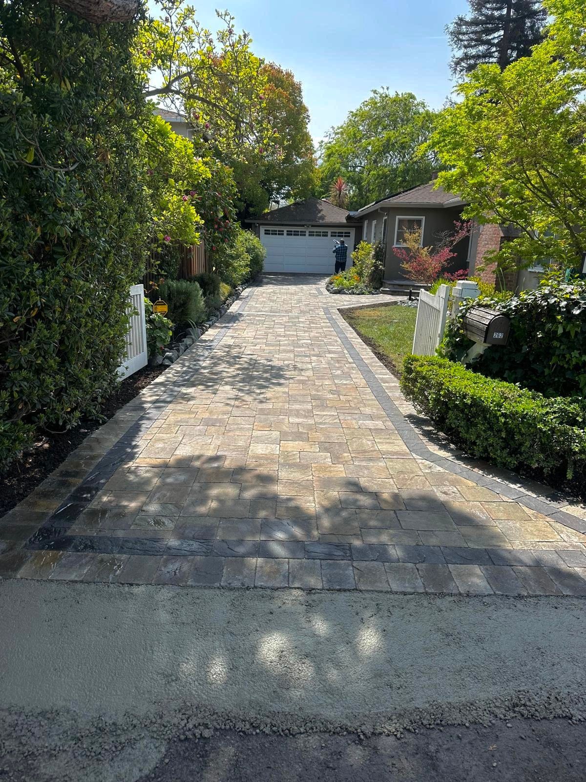 A newly installed stone paver driveway with a dark border, leading to a house with a garage, surrounded by green trees.