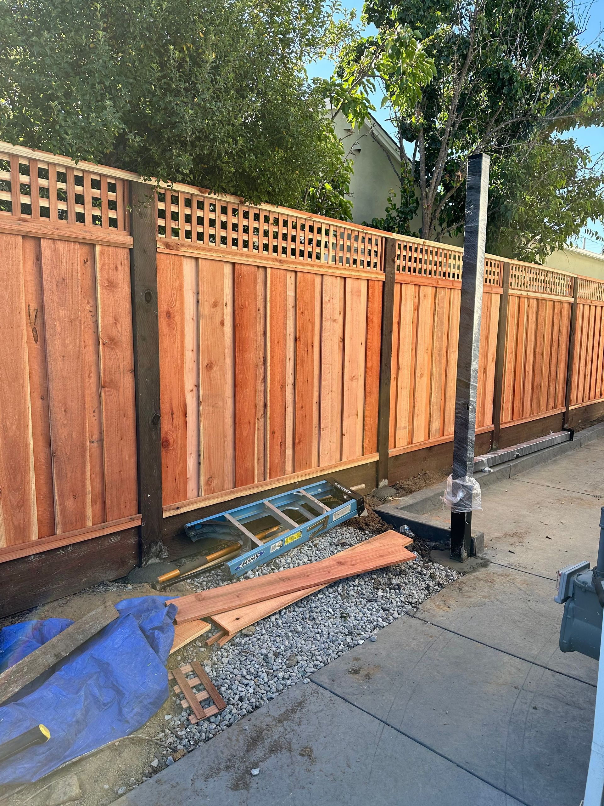 Wooden fence beside a sidewalk with scattered debris and a blue tarp under trees