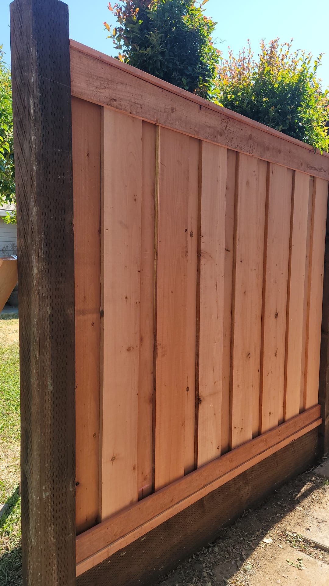 A close-up view of a new cedar fence section with vertical planks, a top cap, and a dark wooden baseboard.
