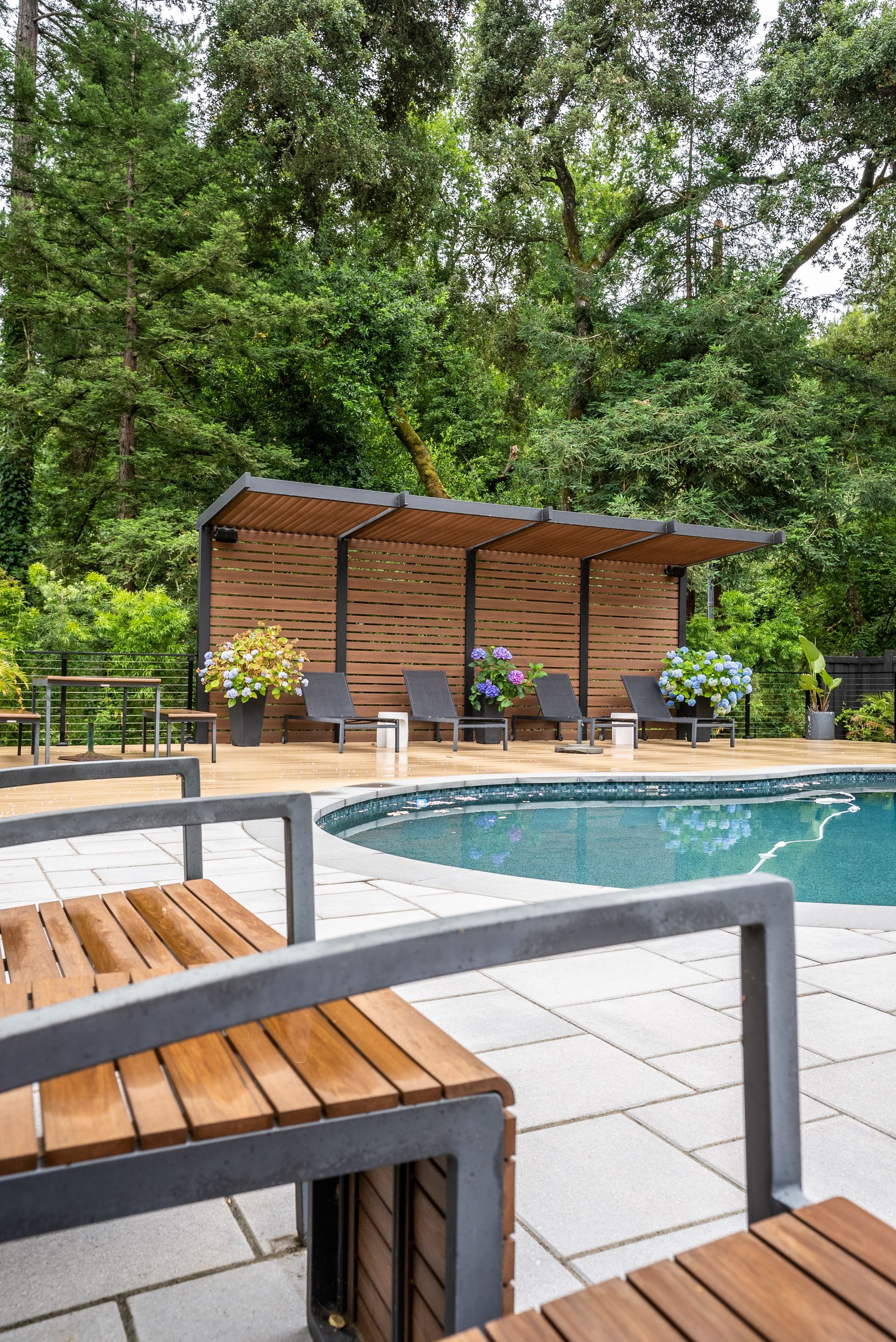Poolside patio with wood pergola, lounge chairs, and greenery beside a swimming pool.