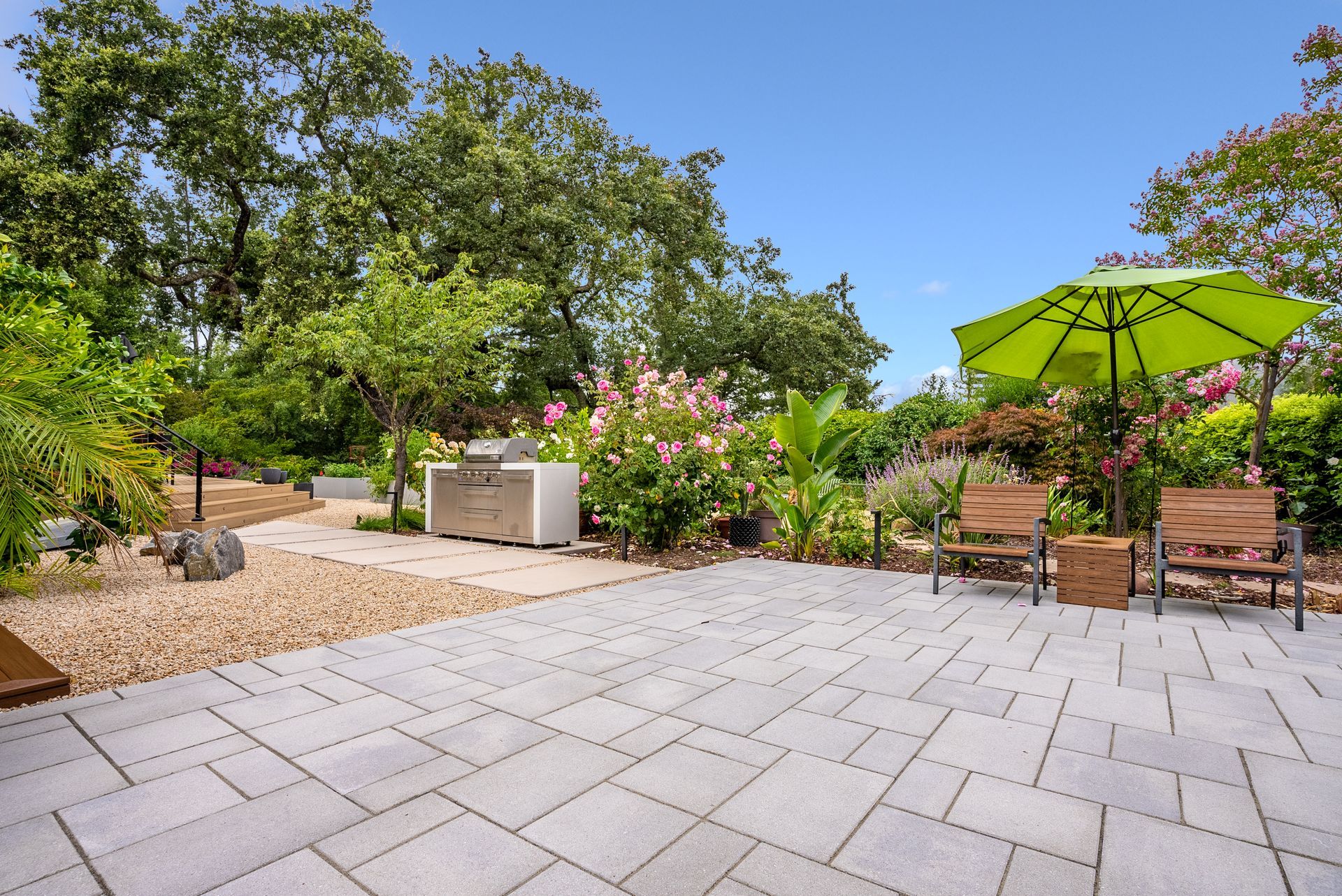 Patio with gray pavers, garden trees and flowers, and a green umbrella shading outdoor chairs and table