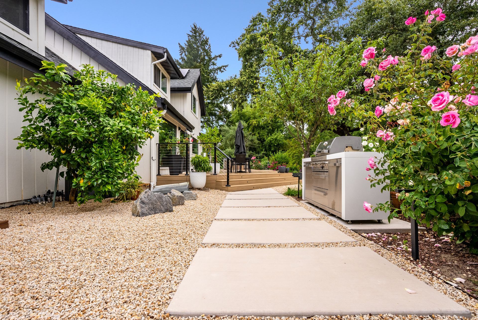 Front walkway beside a house, with gravel, greenery, and pink flowering bushes along the path