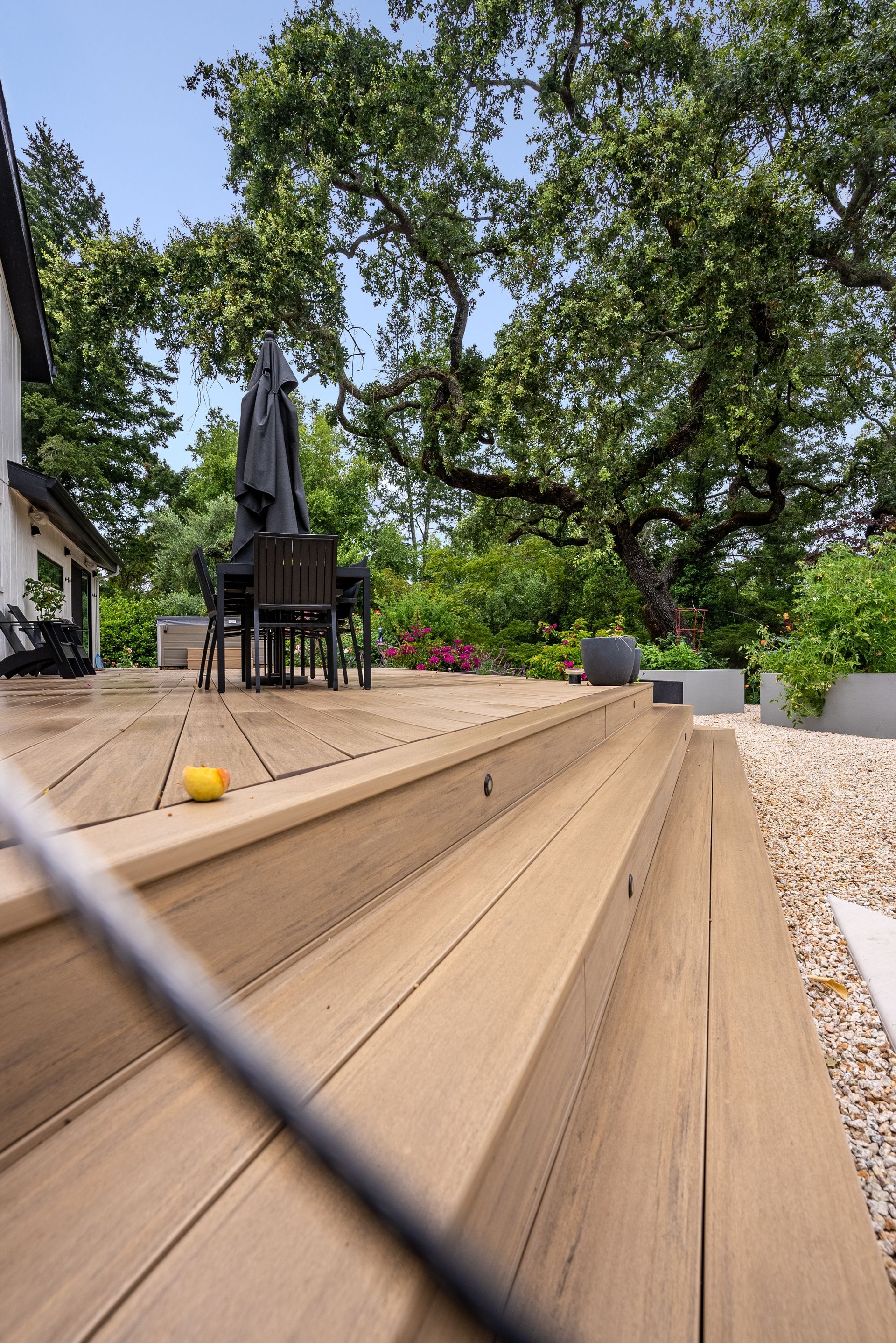 Wooden deck steps leading to a patio with a table, chairs, and a large tree in a backyard.