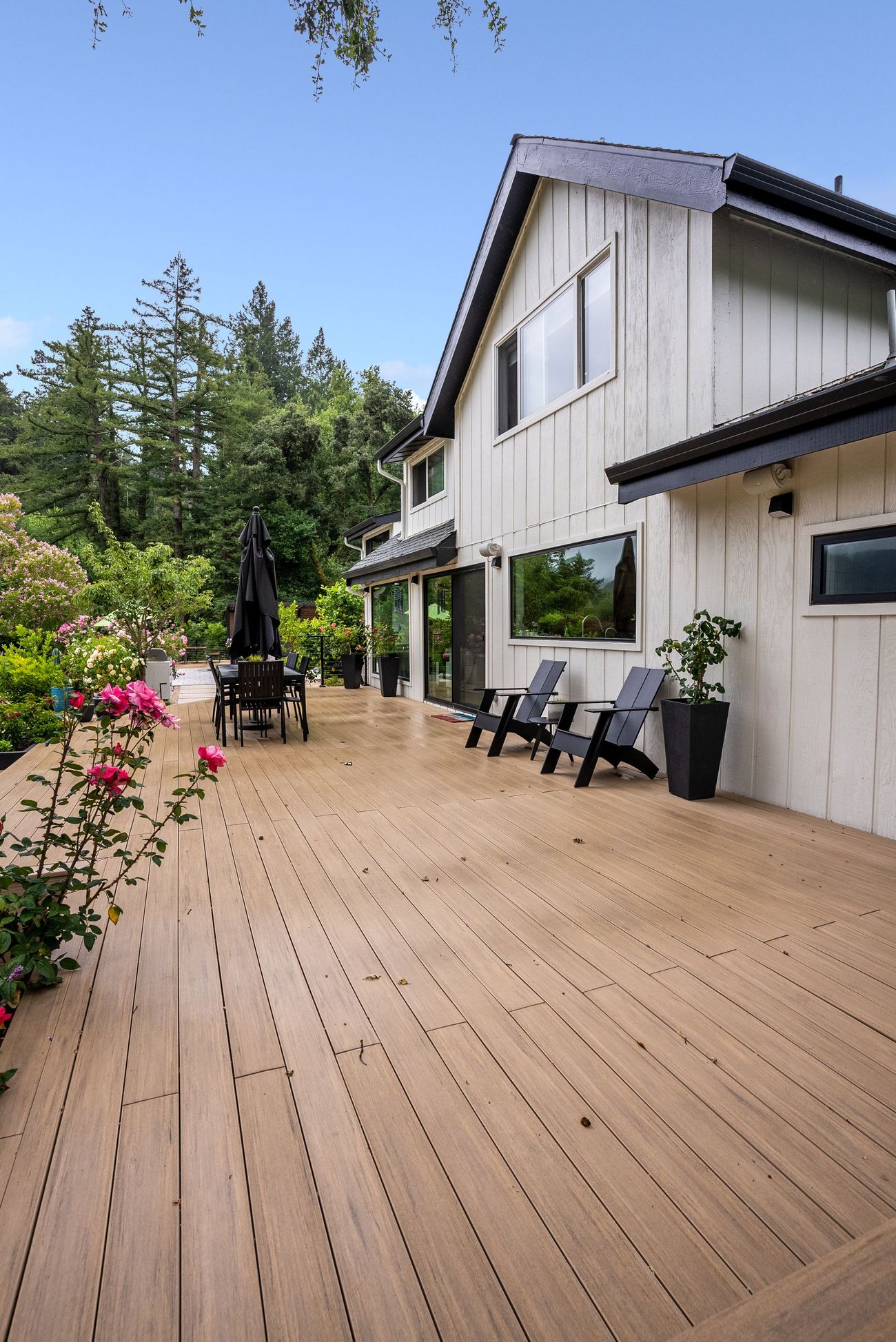 Wooden deck beside a white house, with patio chairs, potted flowers, and trees under a clear blue sky