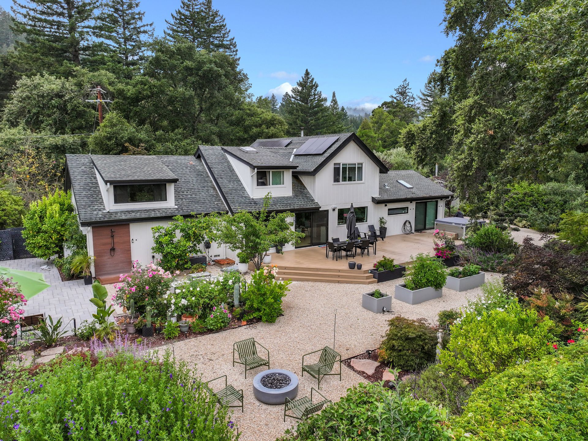 Large house with gray roof, patio, and flower-filled gravel garden surrounded by lush green trees