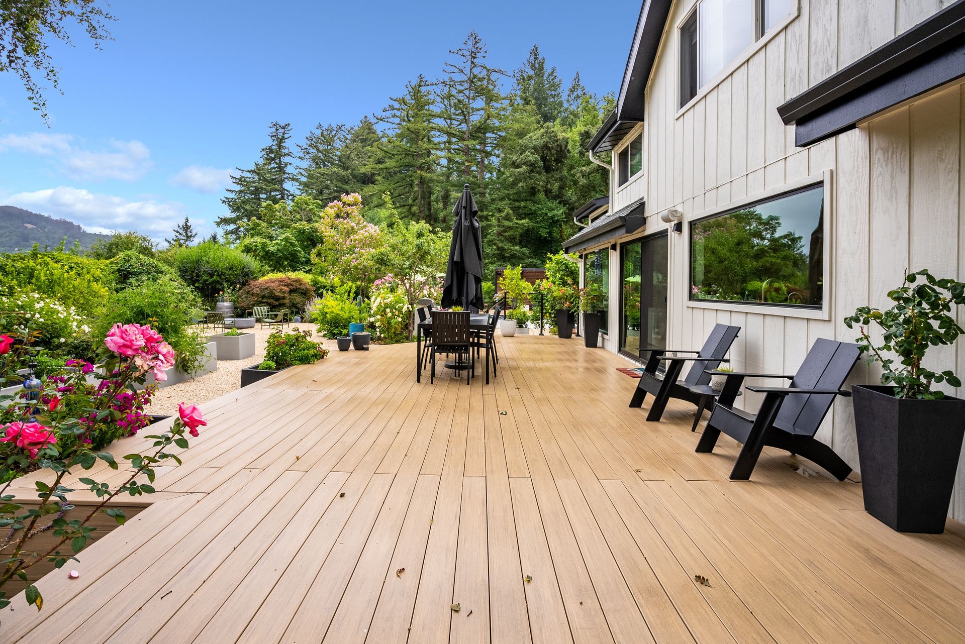 Spacious wooden deck with patio chairs, umbrella, flowers, and mountain views beside a house.