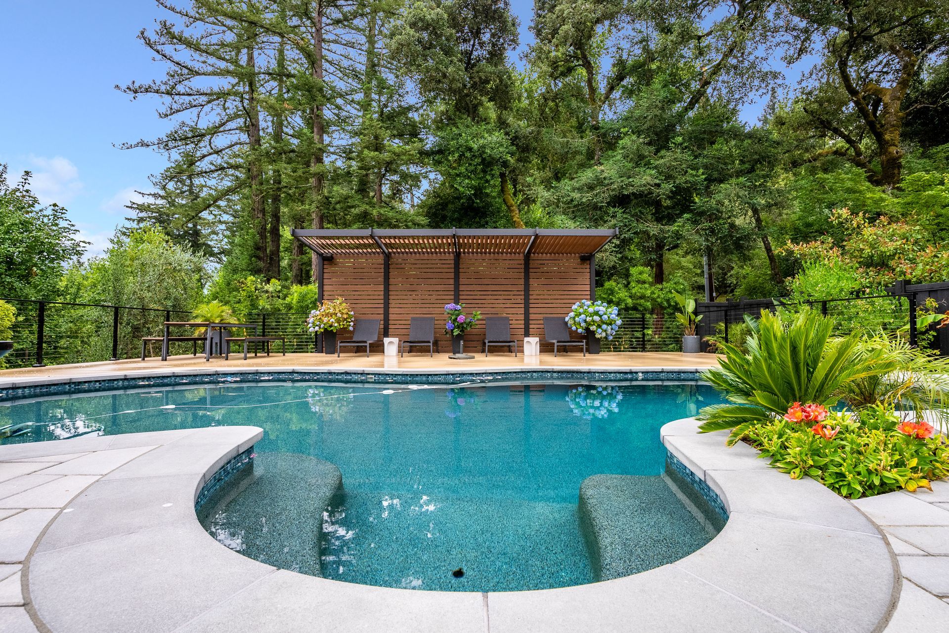 Backyard pool with curved hot tub, stone deck, and lush trees and flowers