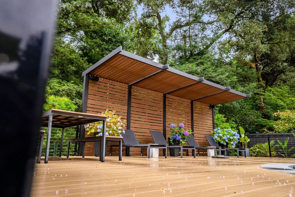 Modern wooden patio shelter with lounge chairs and purple flowers in a lush garden setting