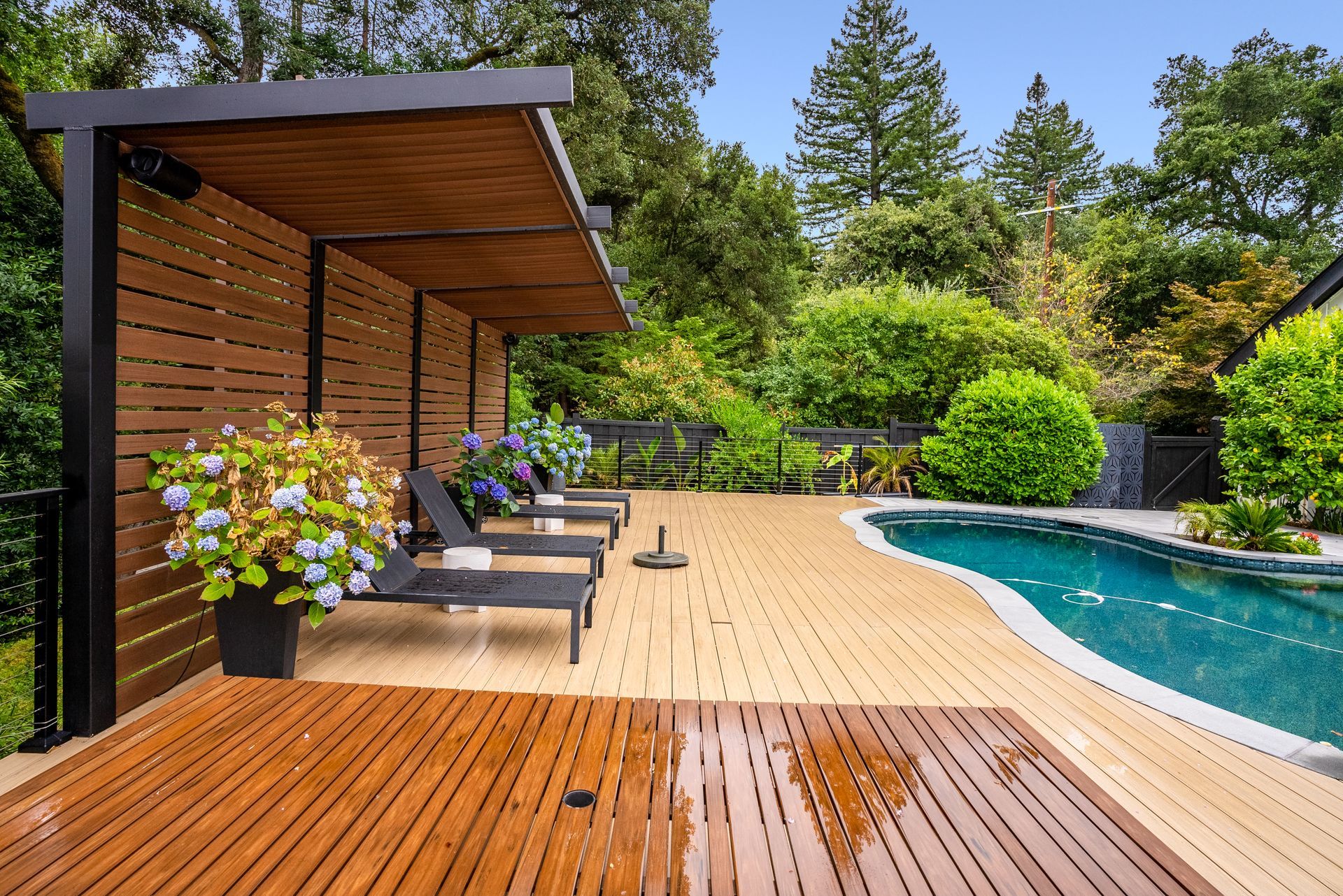 Wooden pool deck with lounge chairs, potted flowers, and a shaded pergola beside a blue swimming pool