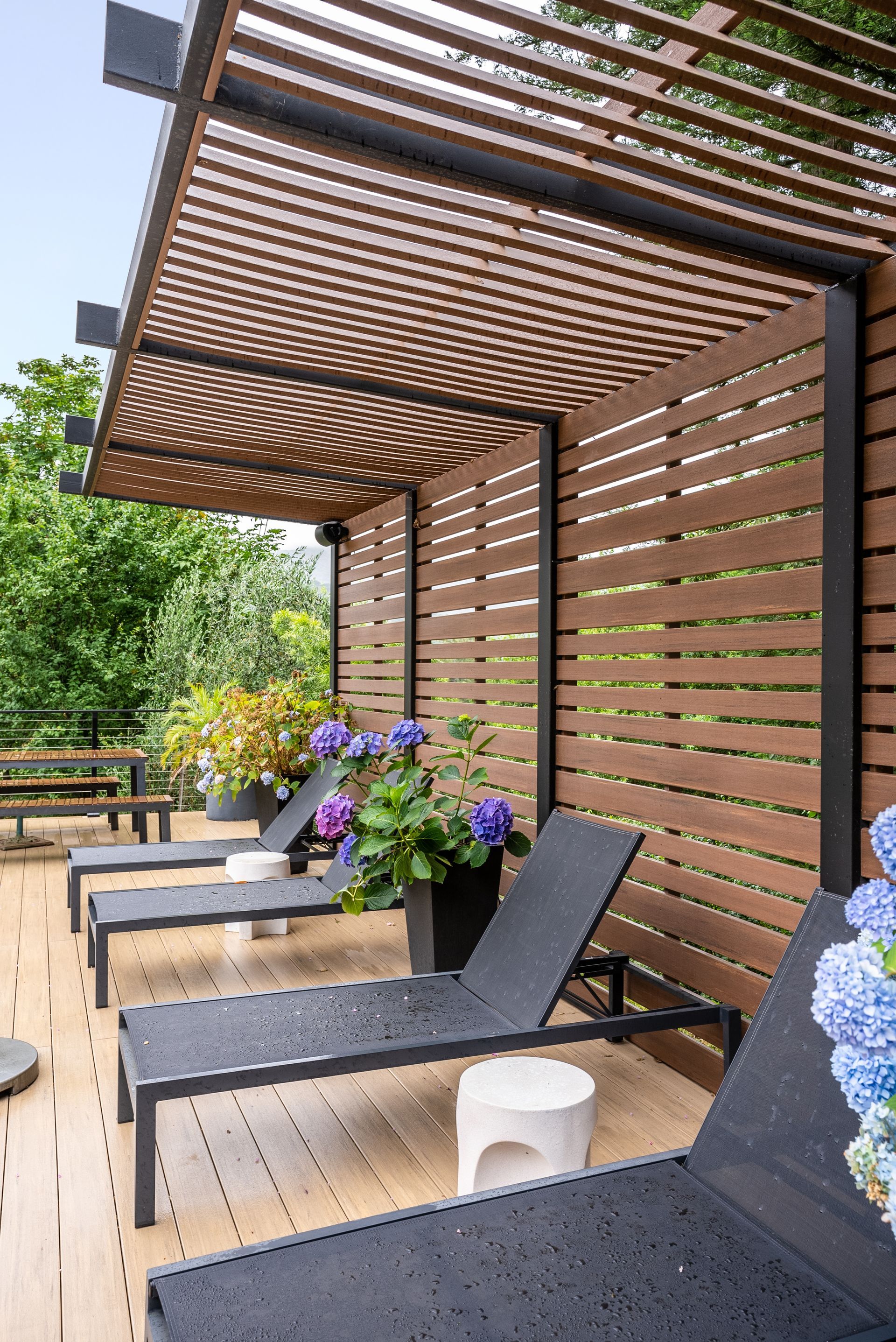 Covered wooden patio with lounge chairs, slatted privacy screen, and purple flowers amid greenery