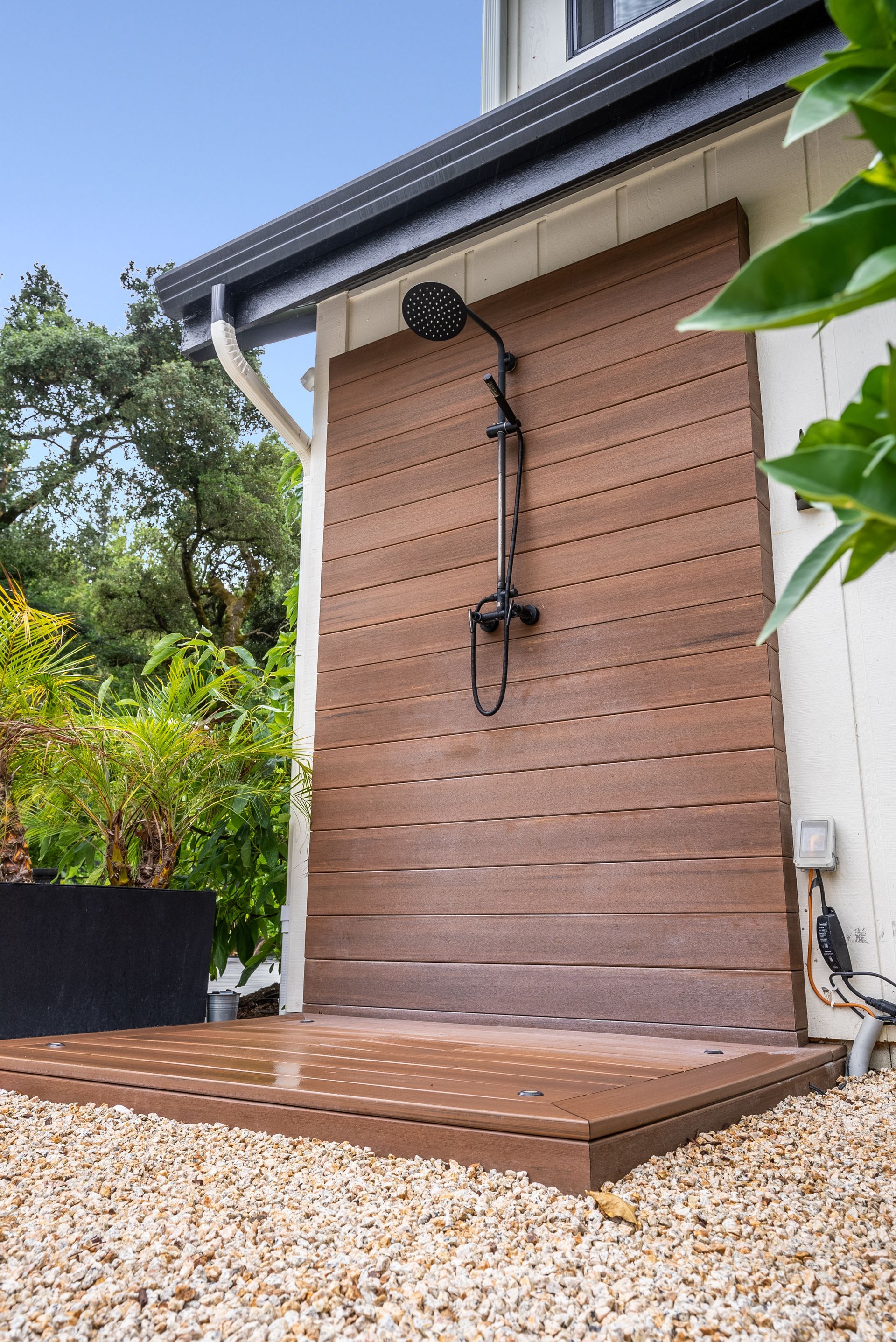 Outdoor wooden shower enclosure beside a house, with a brown slatted door and gravel ground.