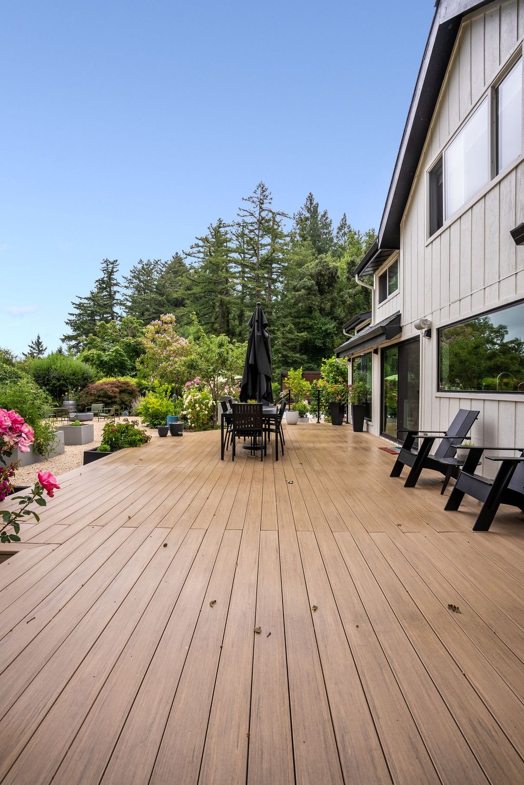 Wooden deck beside a white house, with patio chairs, umbrella, and lush garden greenery.