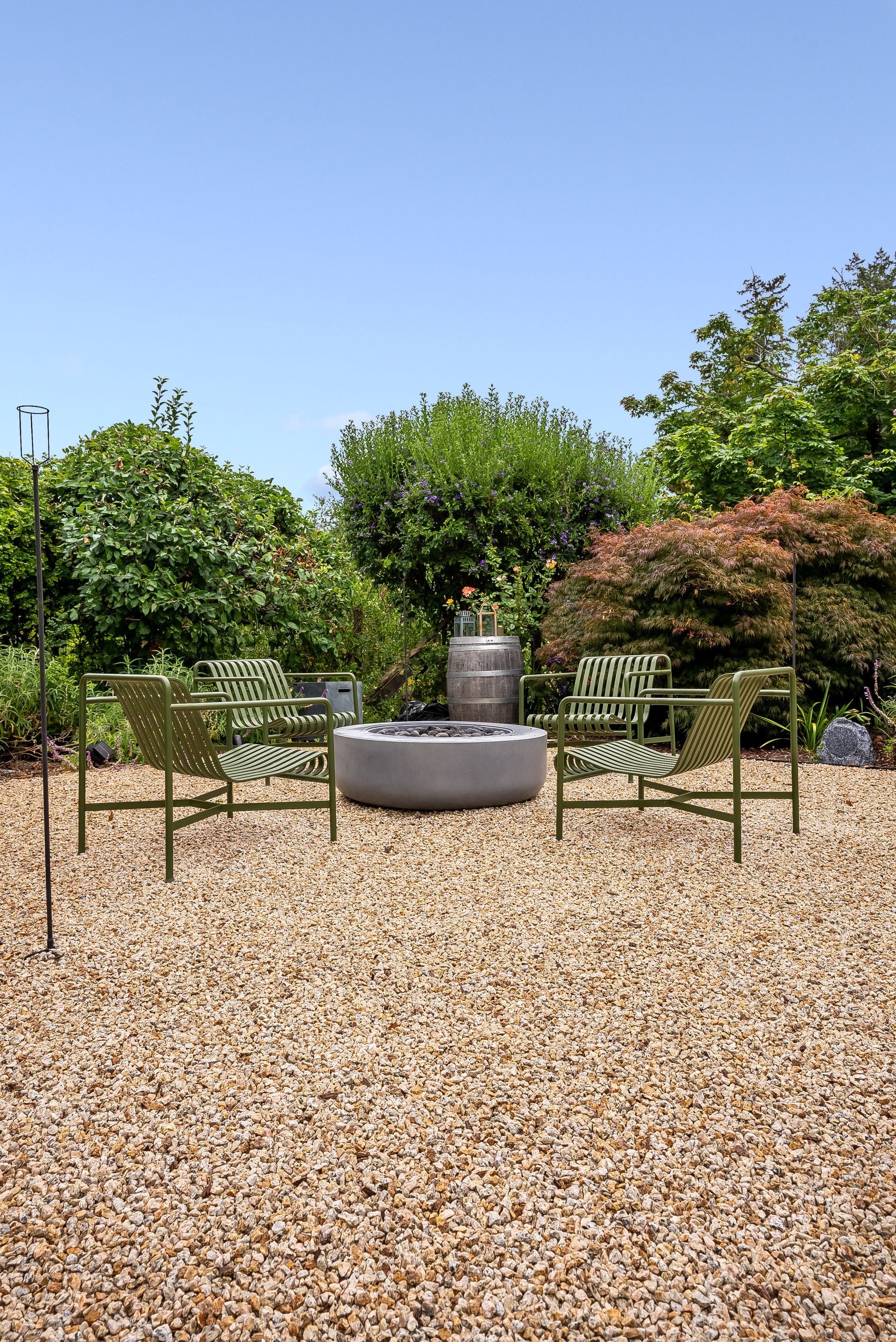 Outdoor patio with two green chairs and a round stone fire pit surrounded by gravel and shrubs under a blue sky