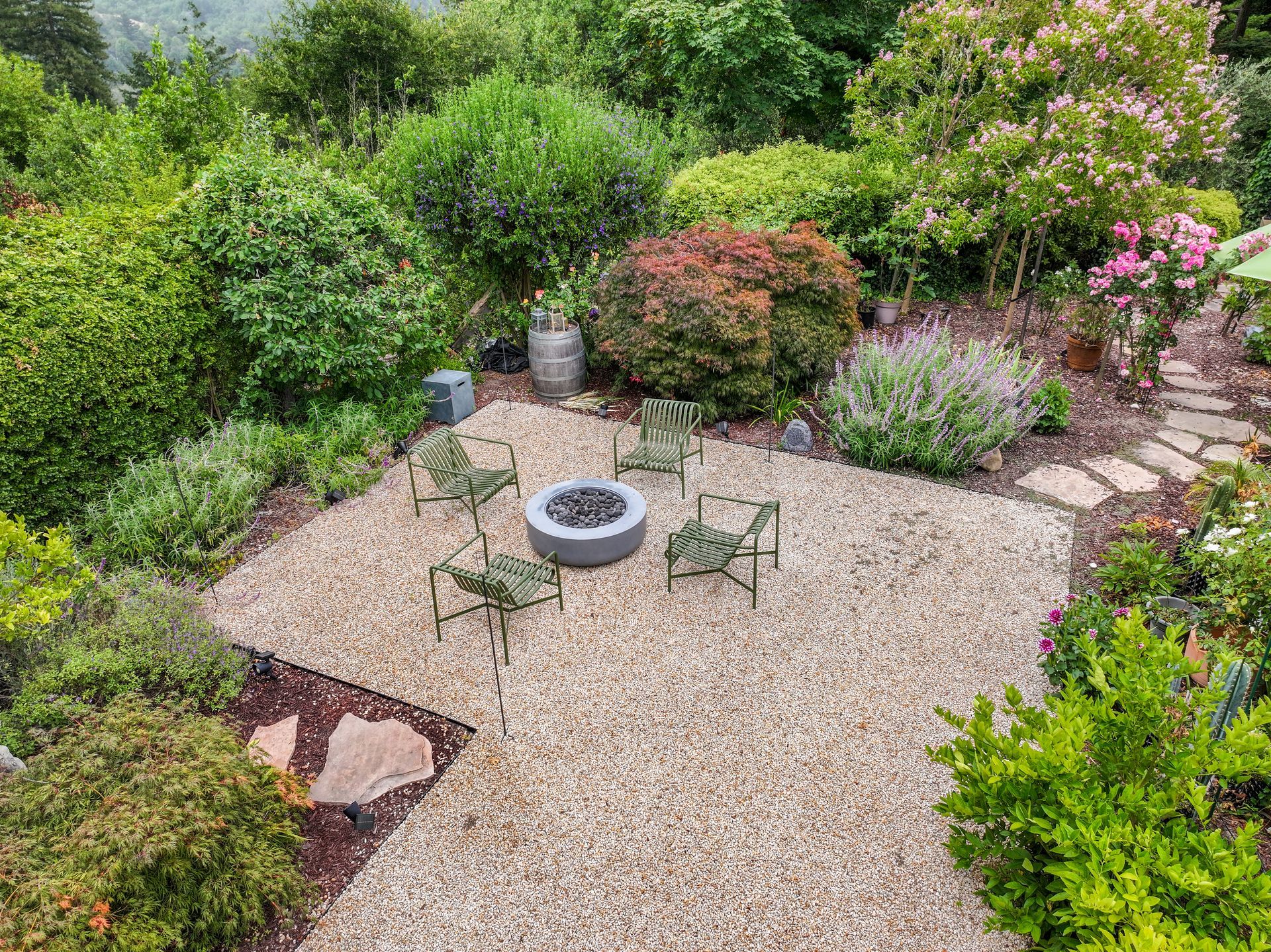 Gravel patio with fire pit and four chairs, surrounded by lush green garden plants and flowers.