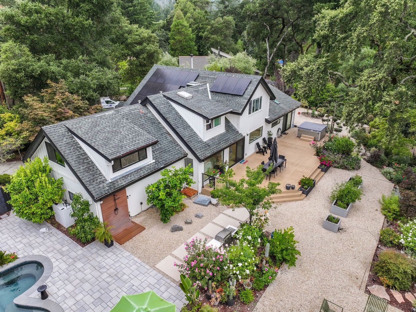 Aerial view of a white house with gray roof, patio, and landscaped garden surrounded by trees